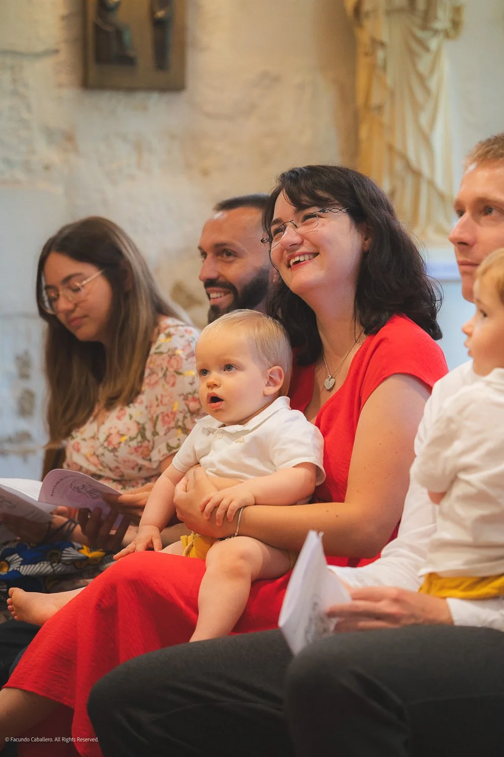 People sitting in a row during a gathering, including a woman wearing a red dress holding a young child, and other adults with notes or programs in hand, in a rustic indoor setting.