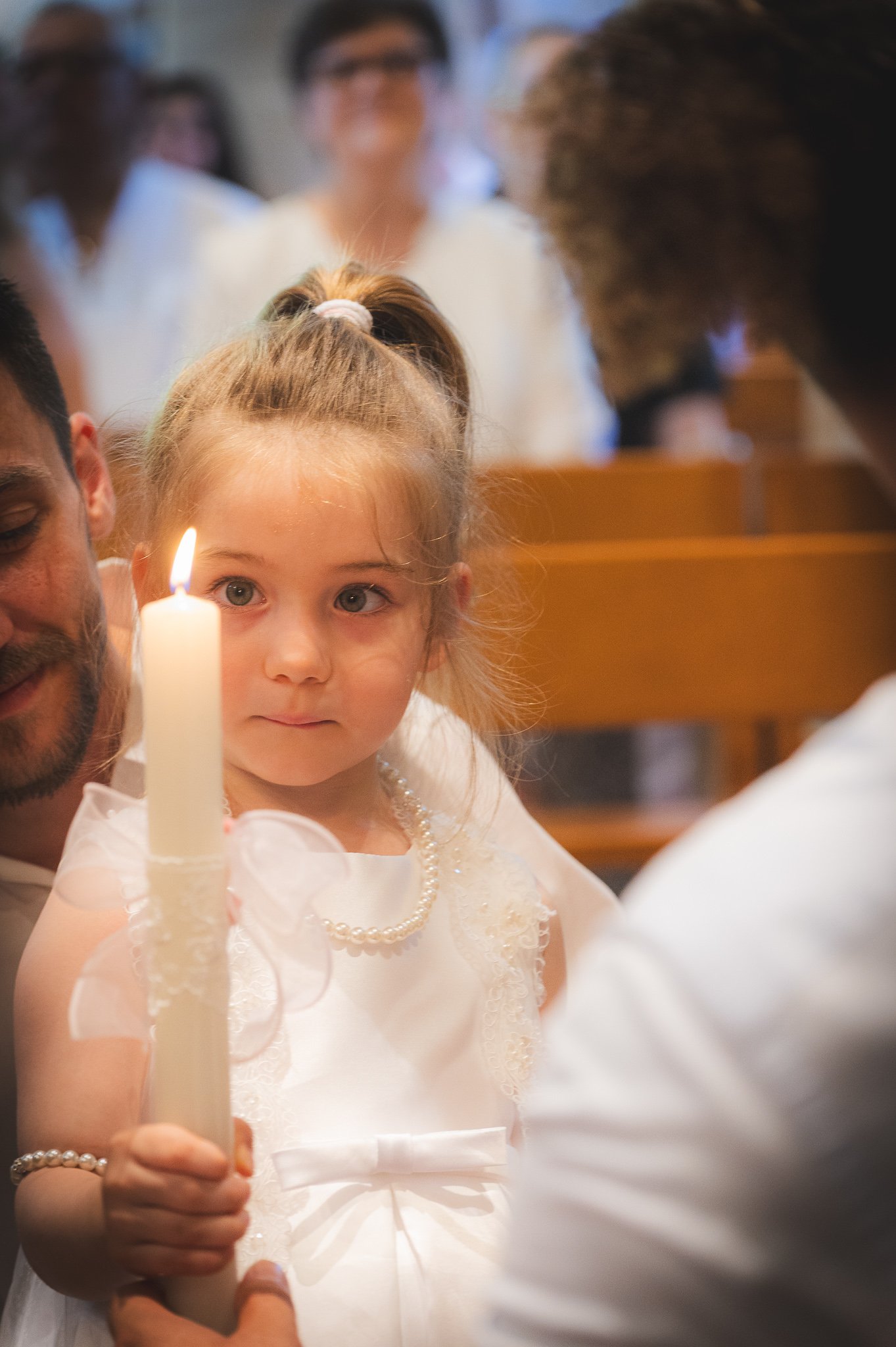 A young girl dressed in white holding a lit candle during a religious ceremony inside a church, with blurred adults in the background.