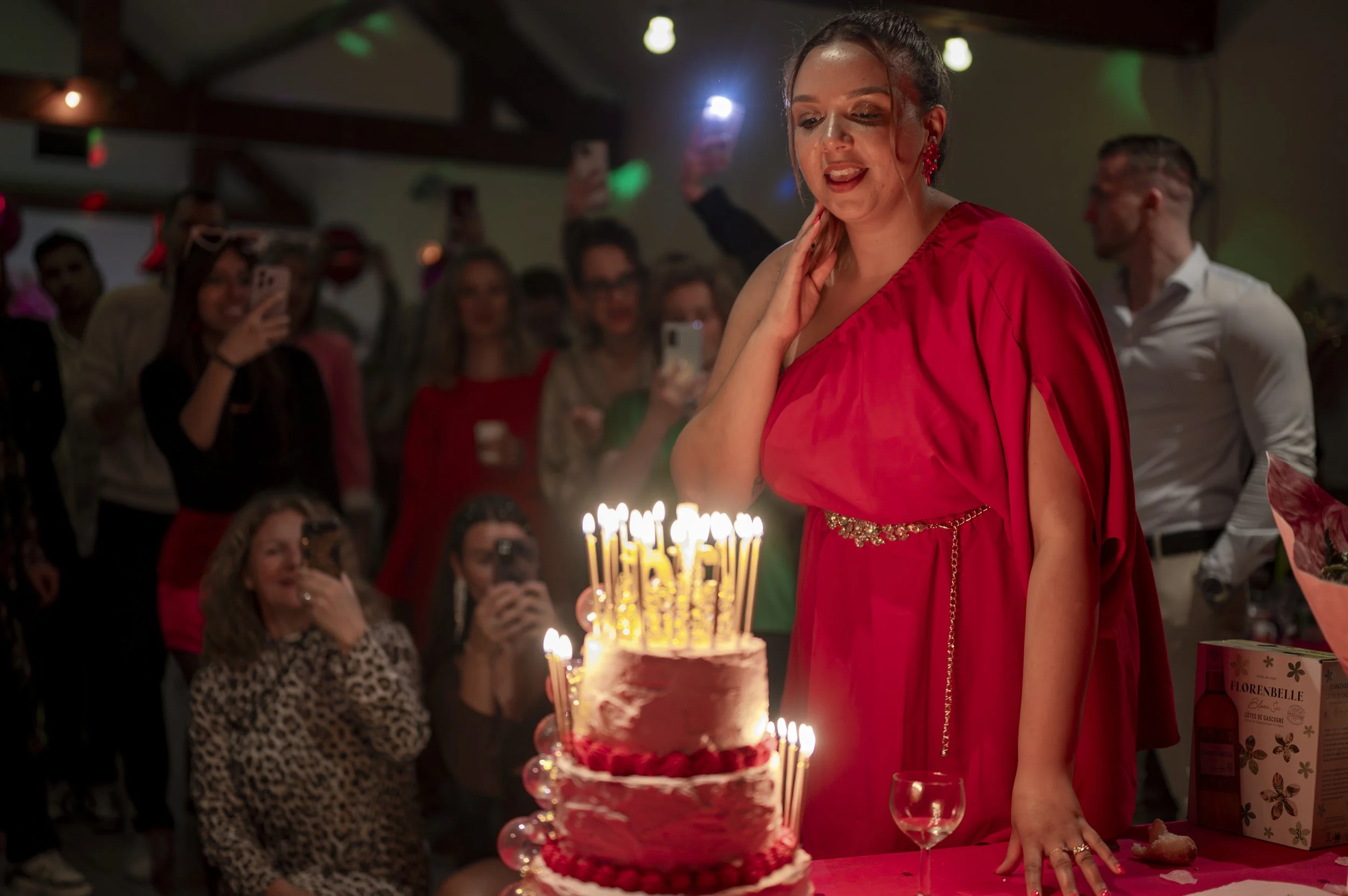 A woman in a red dress stands next to a birthday cake with lit candles at a celebration, surrounded by friends taking photos and watching.