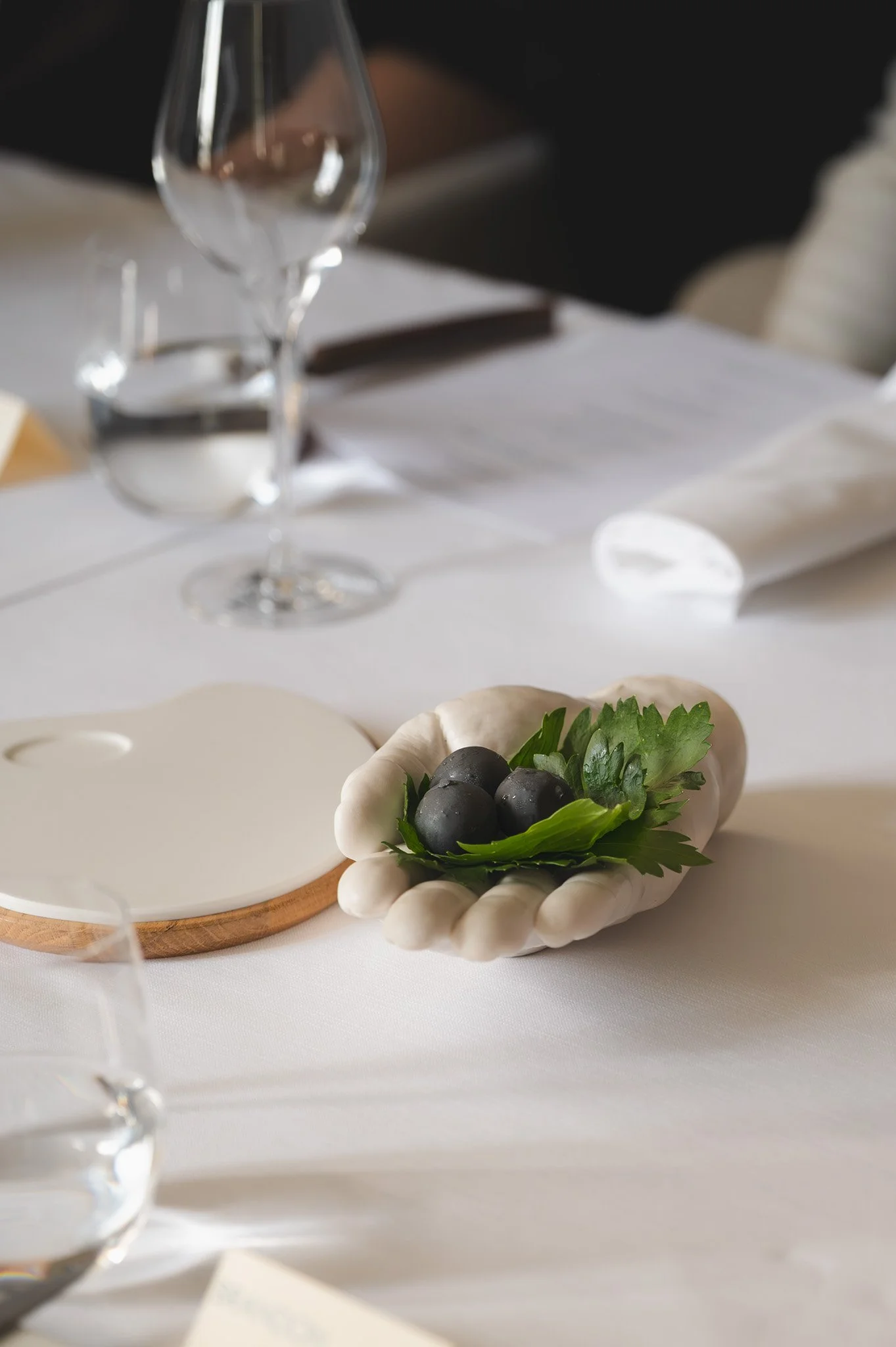 Table setting with a white ceramic bowl containing three dark grapes and green leaves, a wine glass, and a white plate, on a white tablecloth.