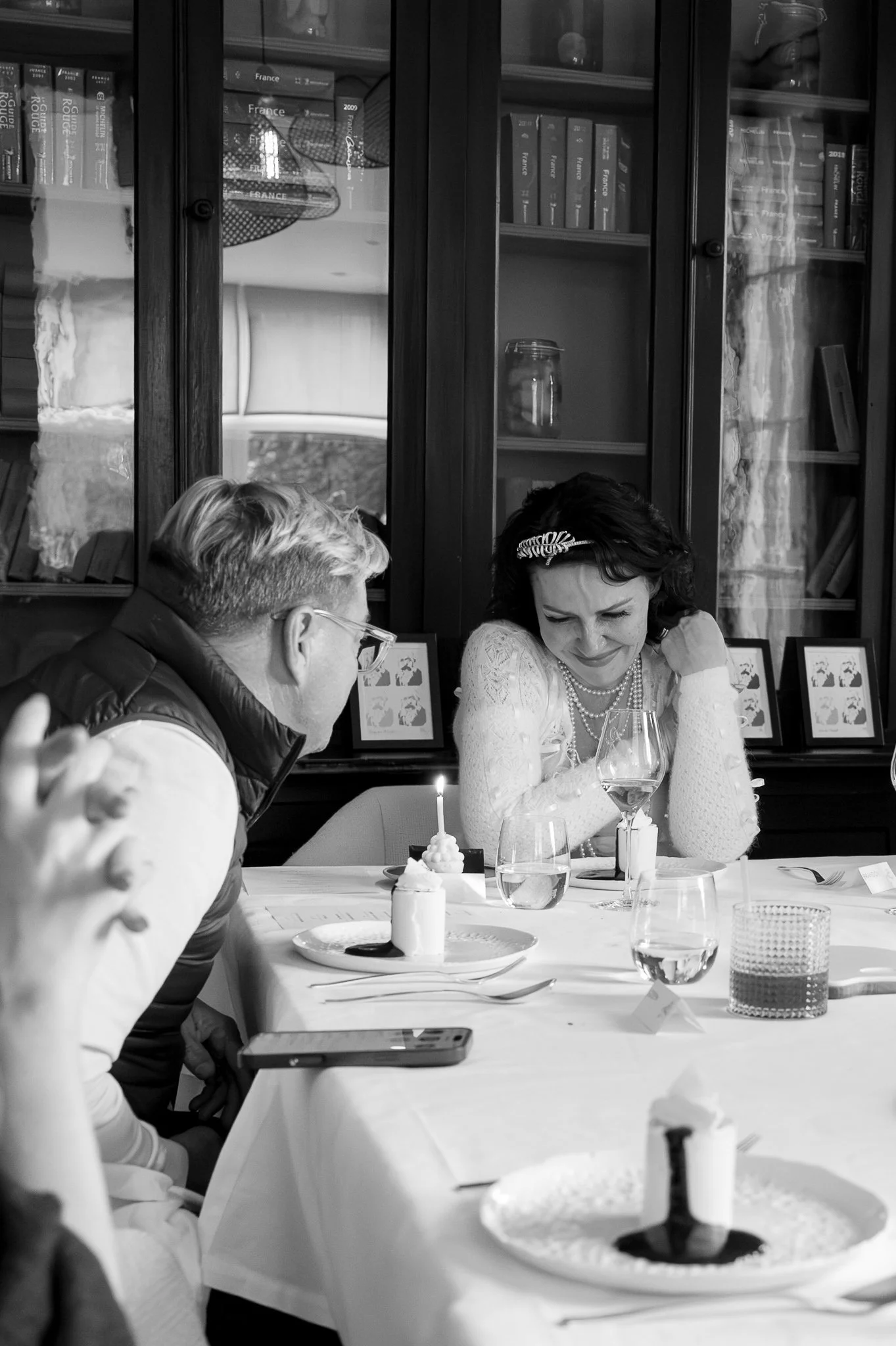 Deux personnes assises à une table lors d'une célébration, avec des desserts et des verres de vin, dans une pièce avec une bibliothèque en arrière-plan.