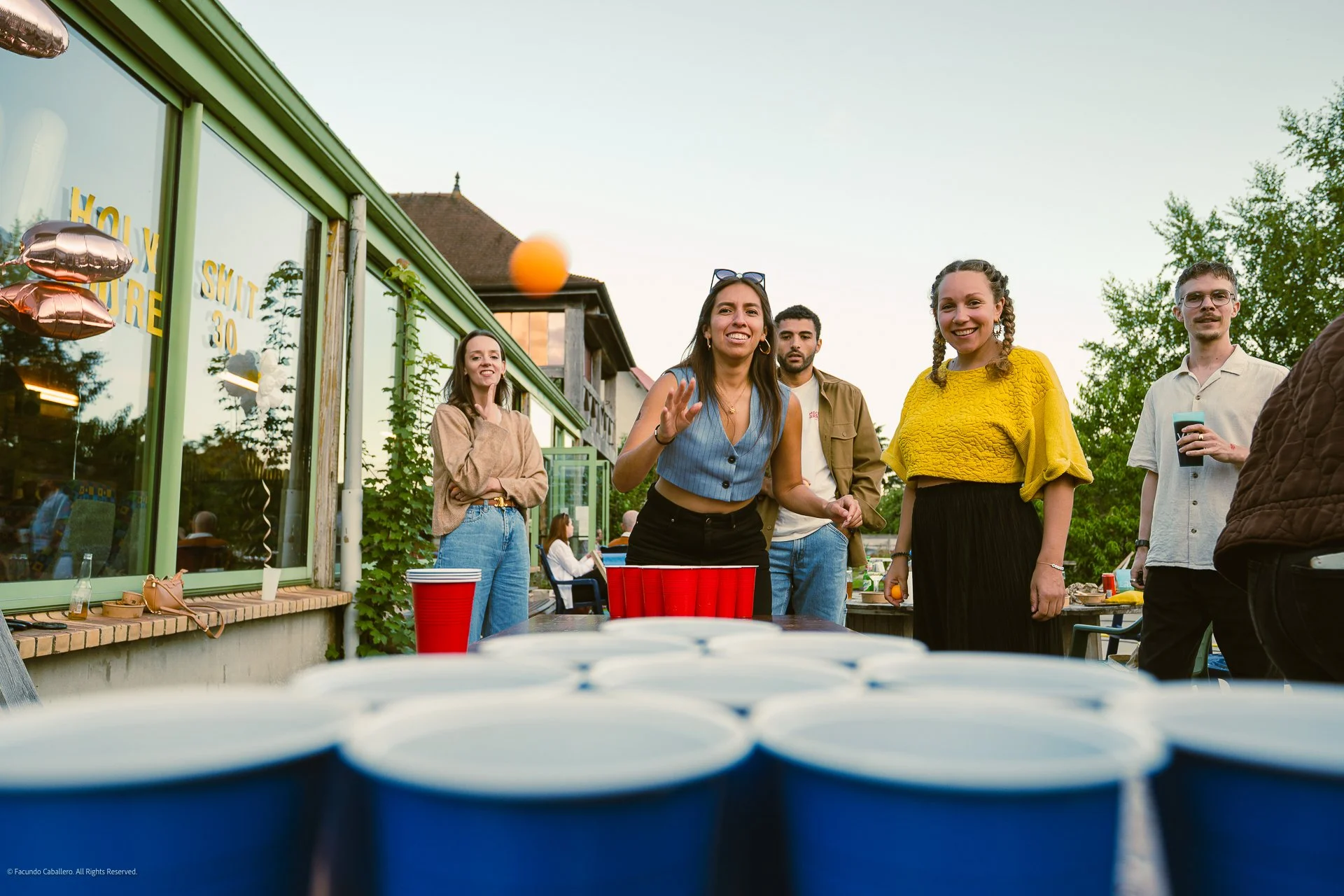 Groupe d'amis jouant au beer pong lors d'une fête en extérieur, avec balles orange et gobelets rouges sur la table.