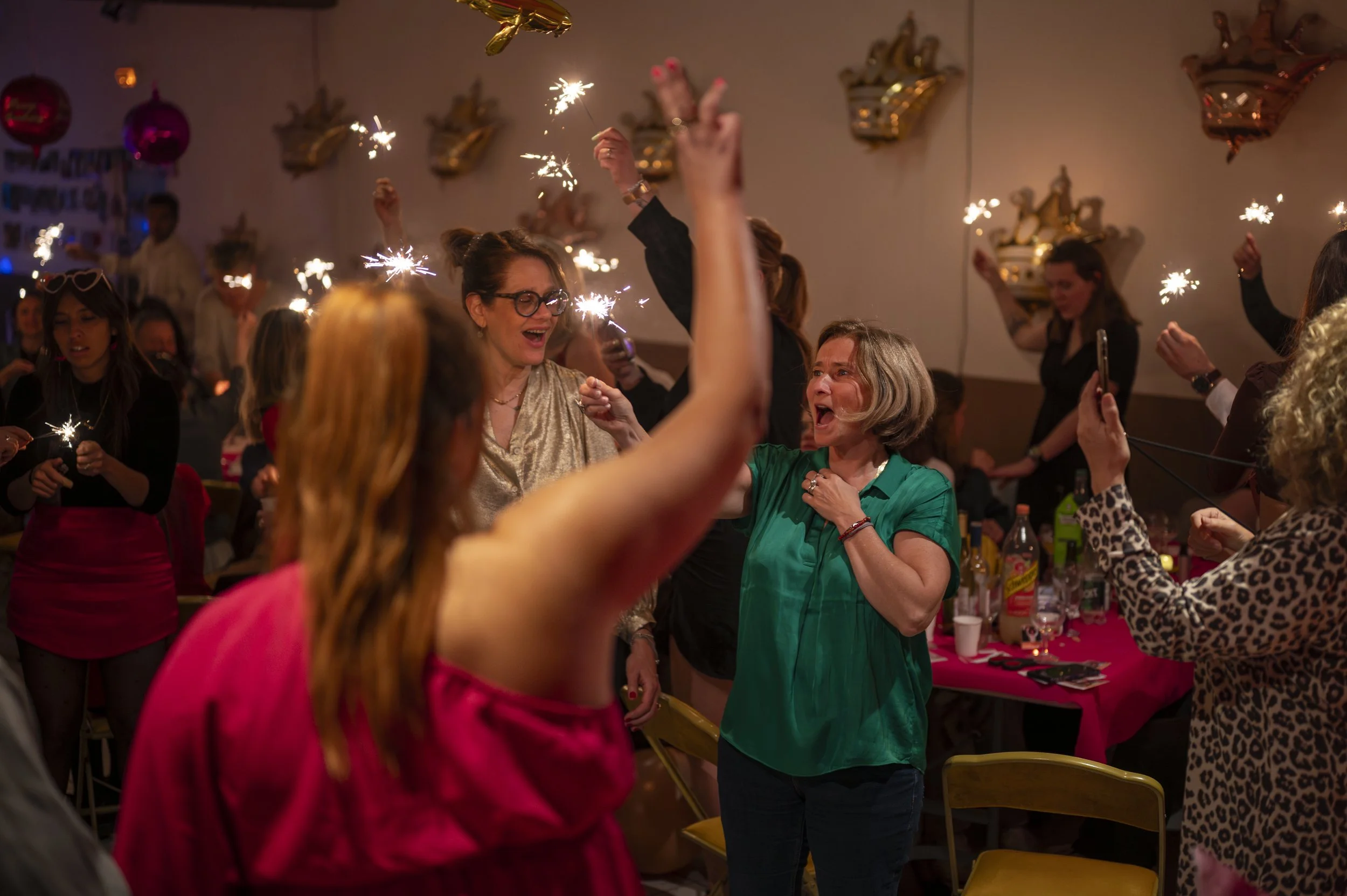 Des femmes souriantes et joyeuses participant à une célébration, utilisant des fusées éclairantes lors d'une fête ou d'un réveillon, avec décorations festives sur le mur.