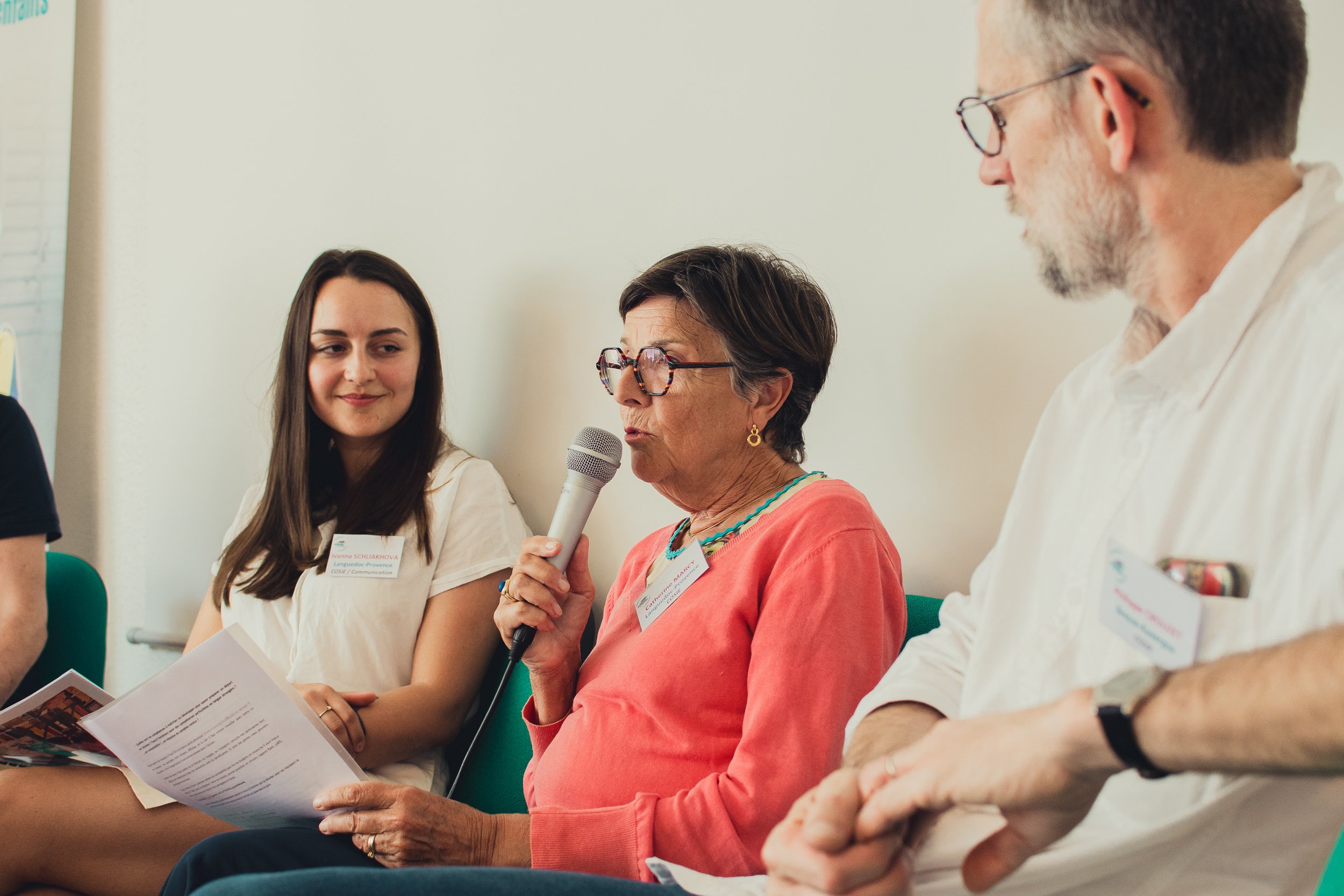 Three people sitting in a panel discussion, woman in the middle speaking into a microphone, two others listening.