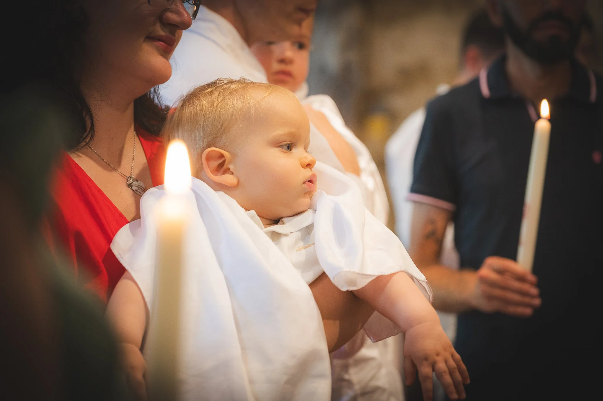 Young boy with blond hair, wearing white, being held by a woman in a red shirt during a religious ceremony, with a candle in front of him. Other people are in the background, some holding candles.