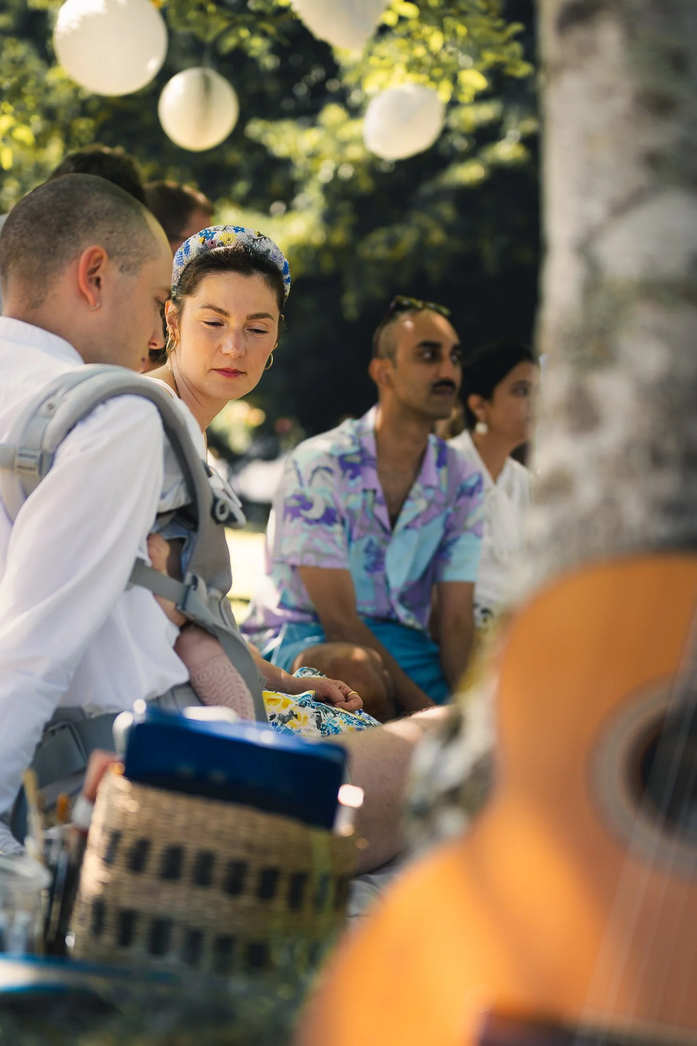 Groupe de personnes assises dehors sous un arbre, en train de jouer de la musique avec une guitare, lors d'un événement ou d'une réunion sociale en journée ensoleillée.