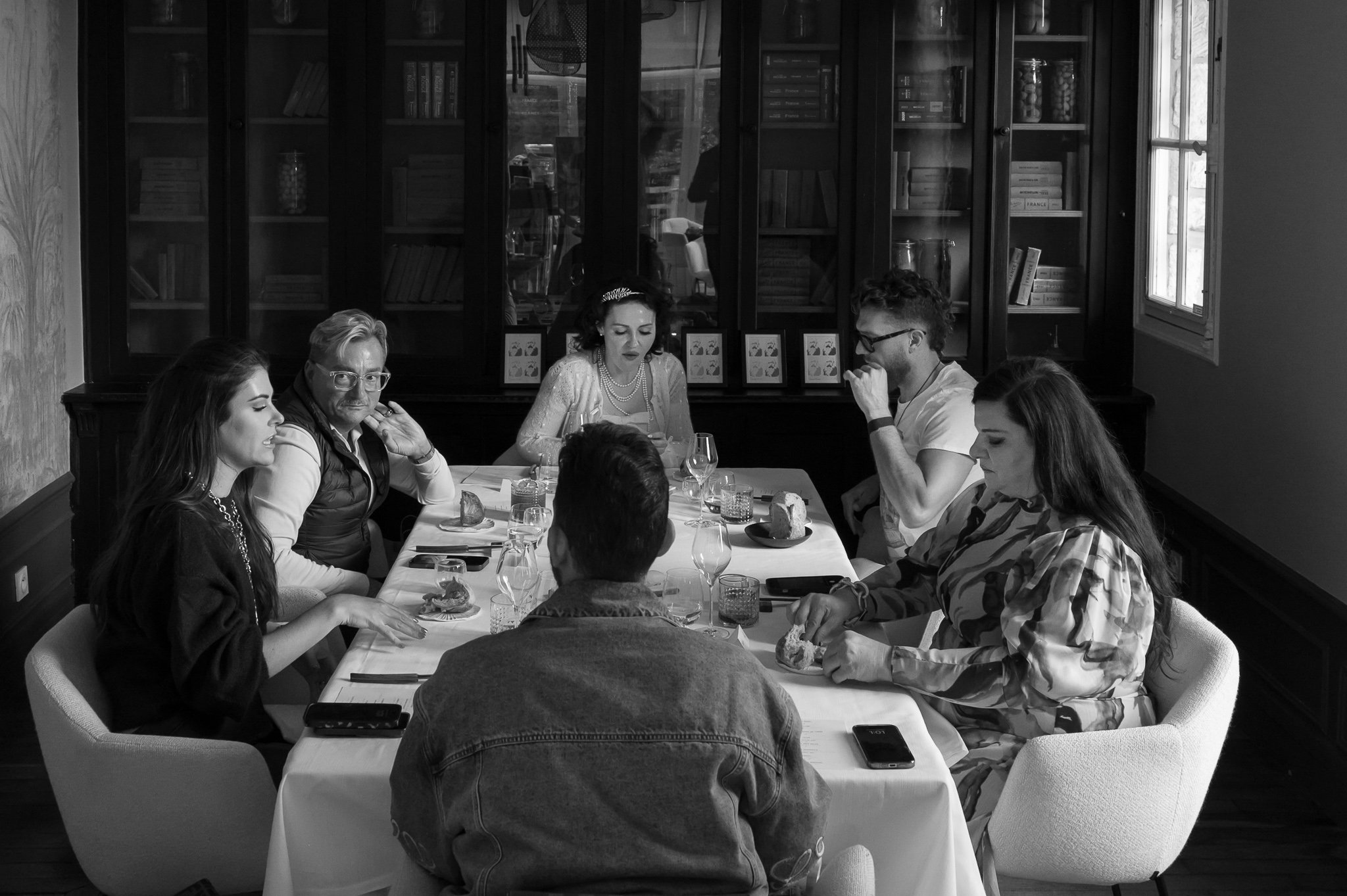 Groupe de sept personnes assises autour d'une table lors d'un repas dans une pièce avec étagères remplies de livres.