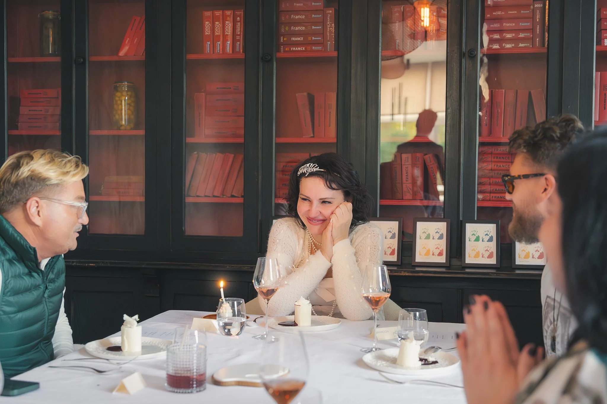 Groupe de personnes lors d'un dîner, une femme au centre souriante avec une couronne de fleurs, entourée de plusieurs amis dans un cadre intérieur avec une armoire noire derrière. La table est dressée avec des verres à vin, des assiettes et des bougi