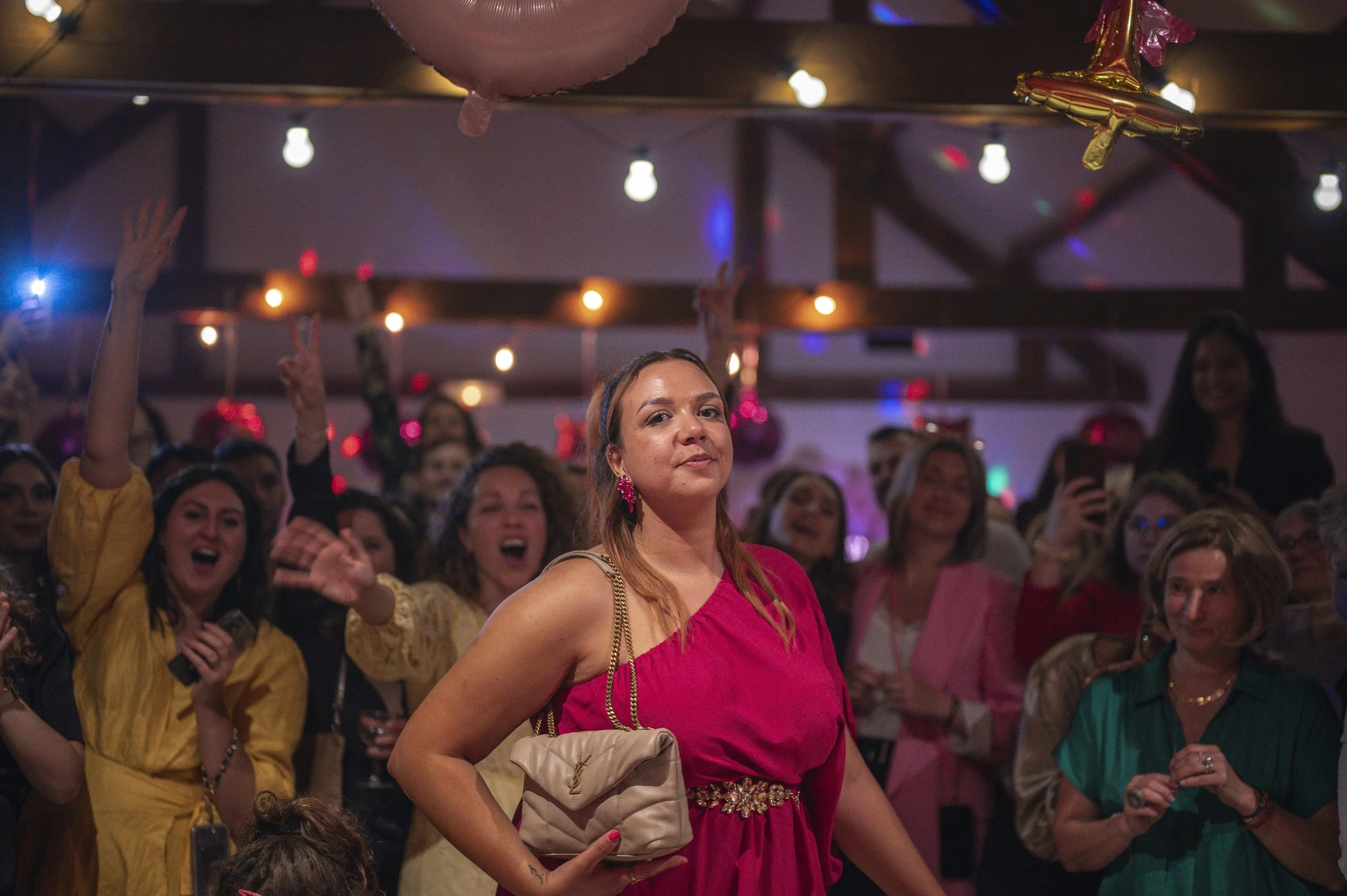 A woman in a pink dress and beige purse stands in front of a crowd at a party or celebration, with people smiling and cheering in the background.