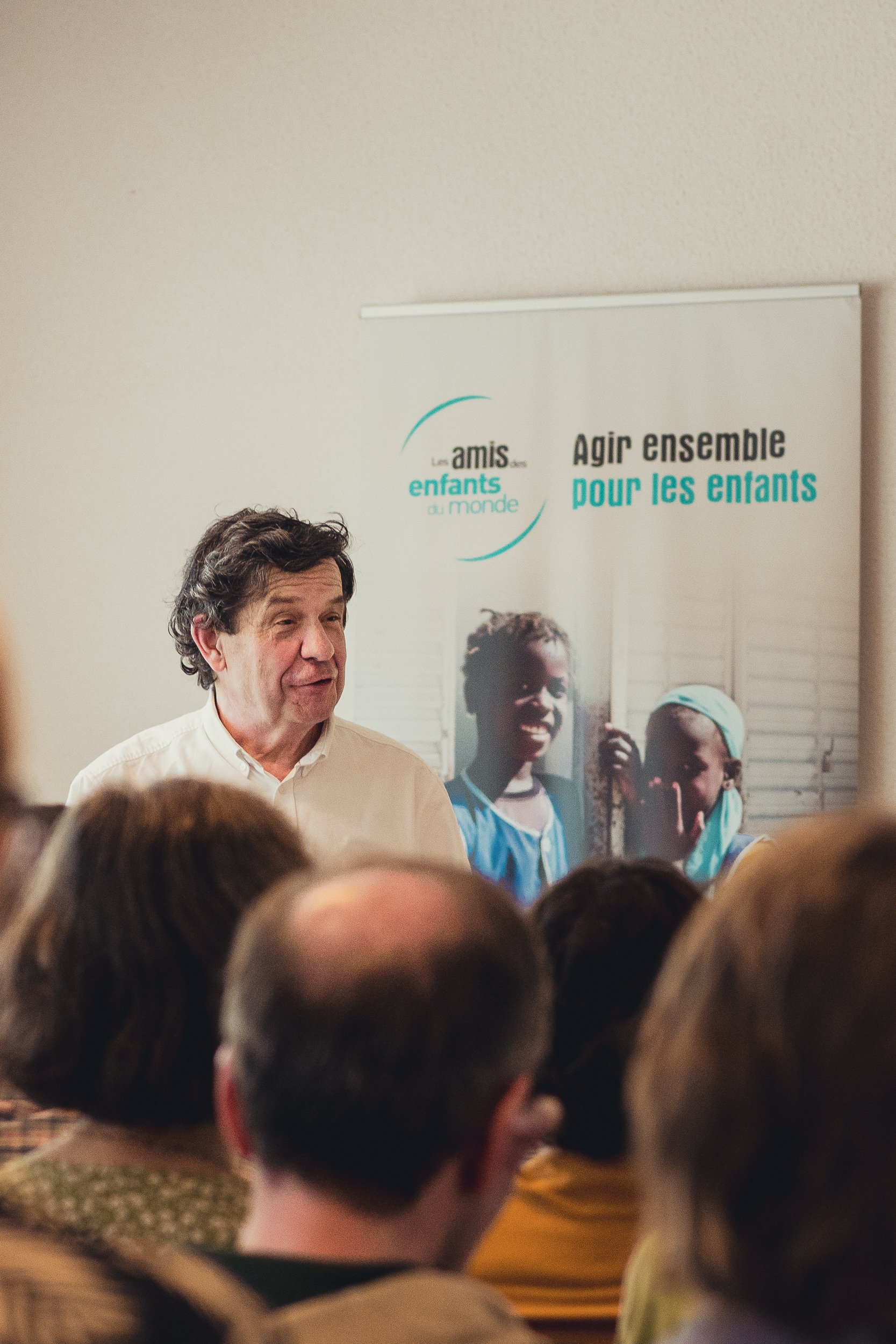 A man speaking at a community event with a poster behind him that reads 'Les amis des enfants du monde, Agir ensemble pour les enfants' and shows children smiling.