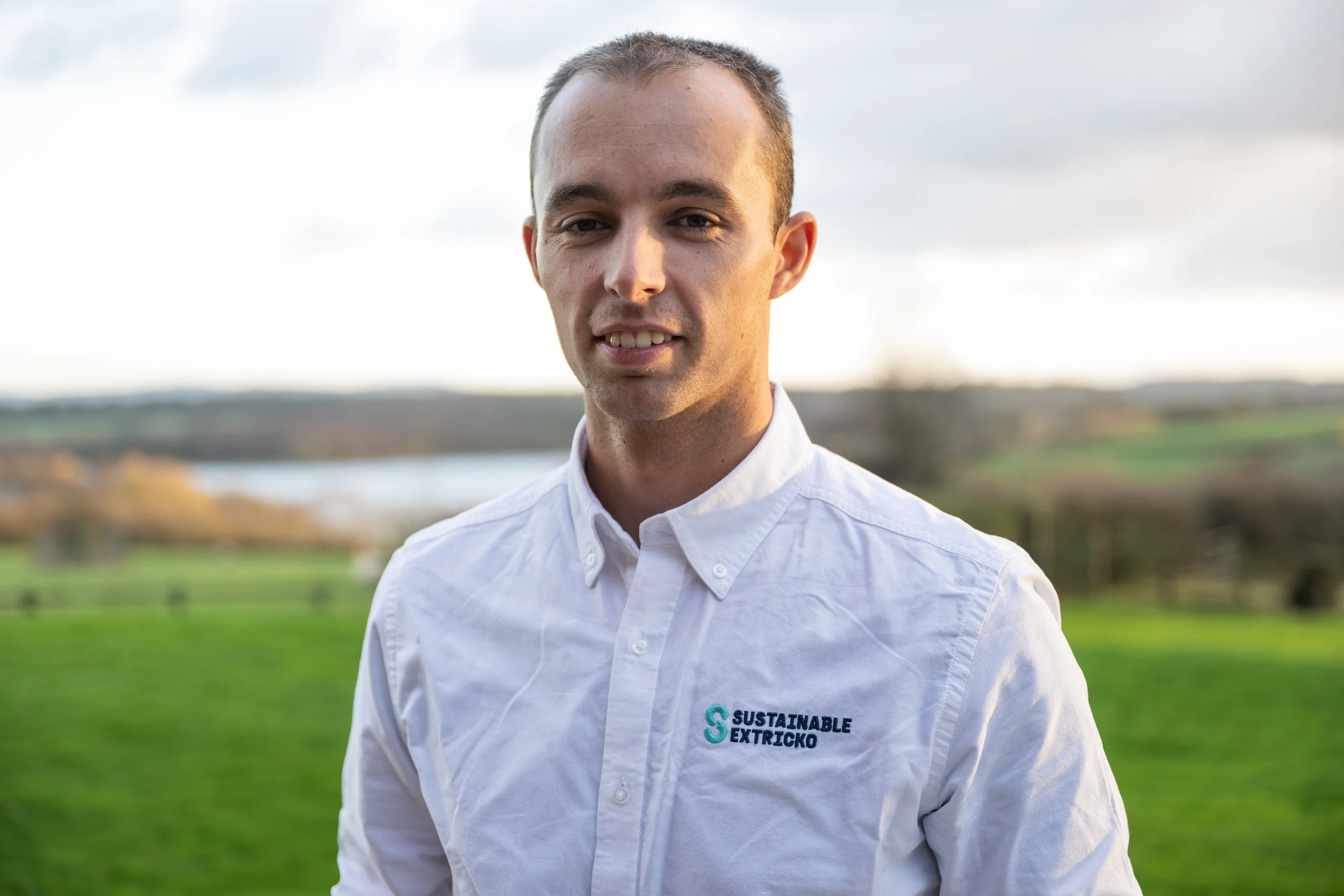 Man in a white shirt with a logo that reads, 'Sustainable Extricko,' standing outdoors with a blurred natural landscape in the background.