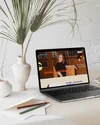 A laptop on a white desk displaying a website titled "Astrology and Lifestyle" with a woman sitting in front of a wooden bookshelf. The desk also has some papers, two colored pencils, and two vases with large palm leaves.