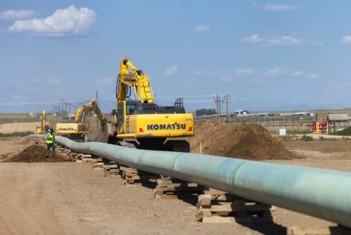 Construction scene with a Komatsu excavator installing a large pipeline on a dirt road, workers in safety vests, clear blue sky with some clouds.