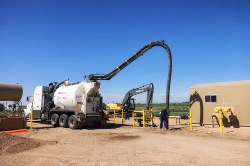 Construction site with vacuum truck, excavator, and workers near portable building under clear blue sky.