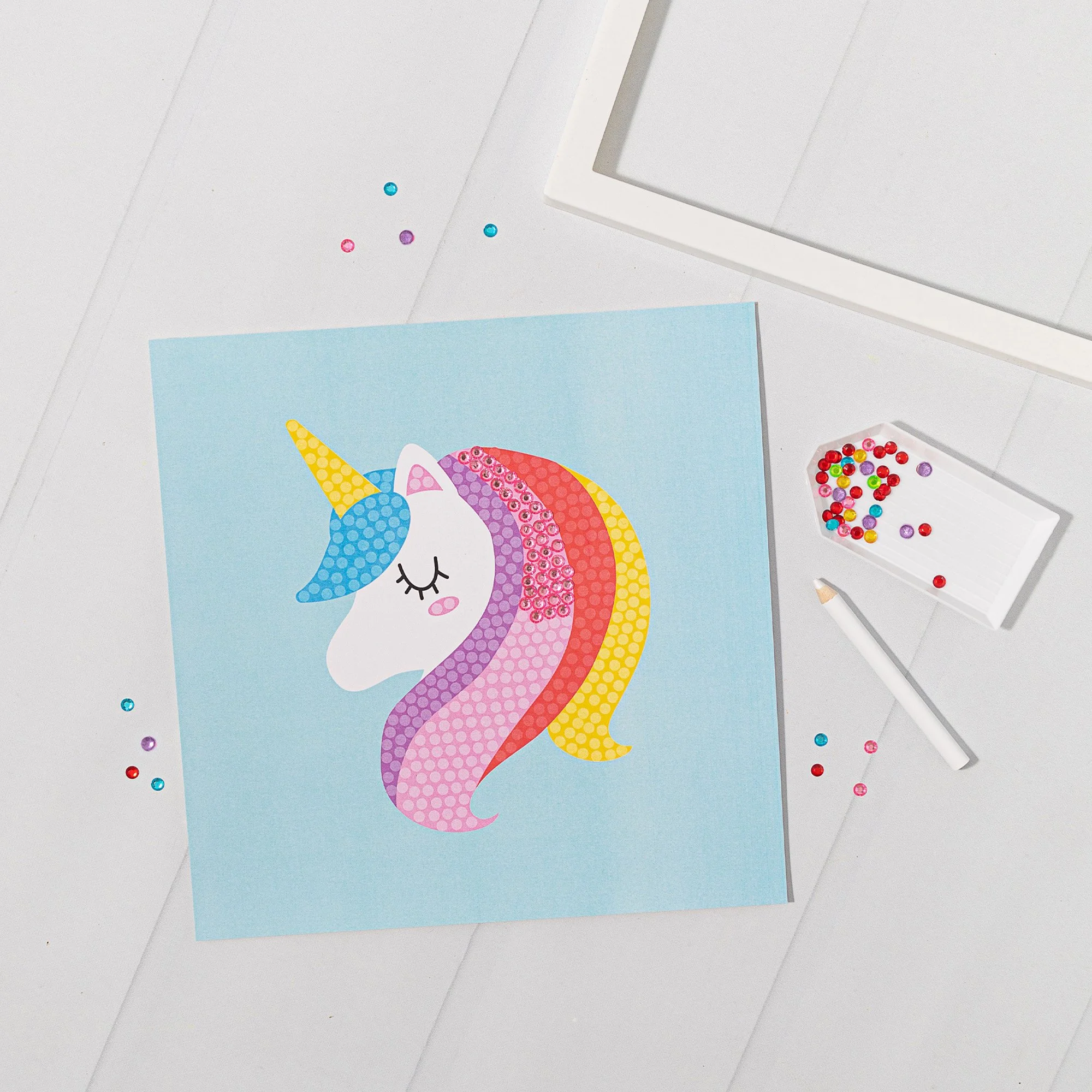 Unicorn craft with pink, purple, red, and yellow rainbow-colored mane and tail, blue horn, and closed eyes, on blue paper. Crafting supplies including beads, a white marker, and a white tray with colorful beads are visible on a white surface.
