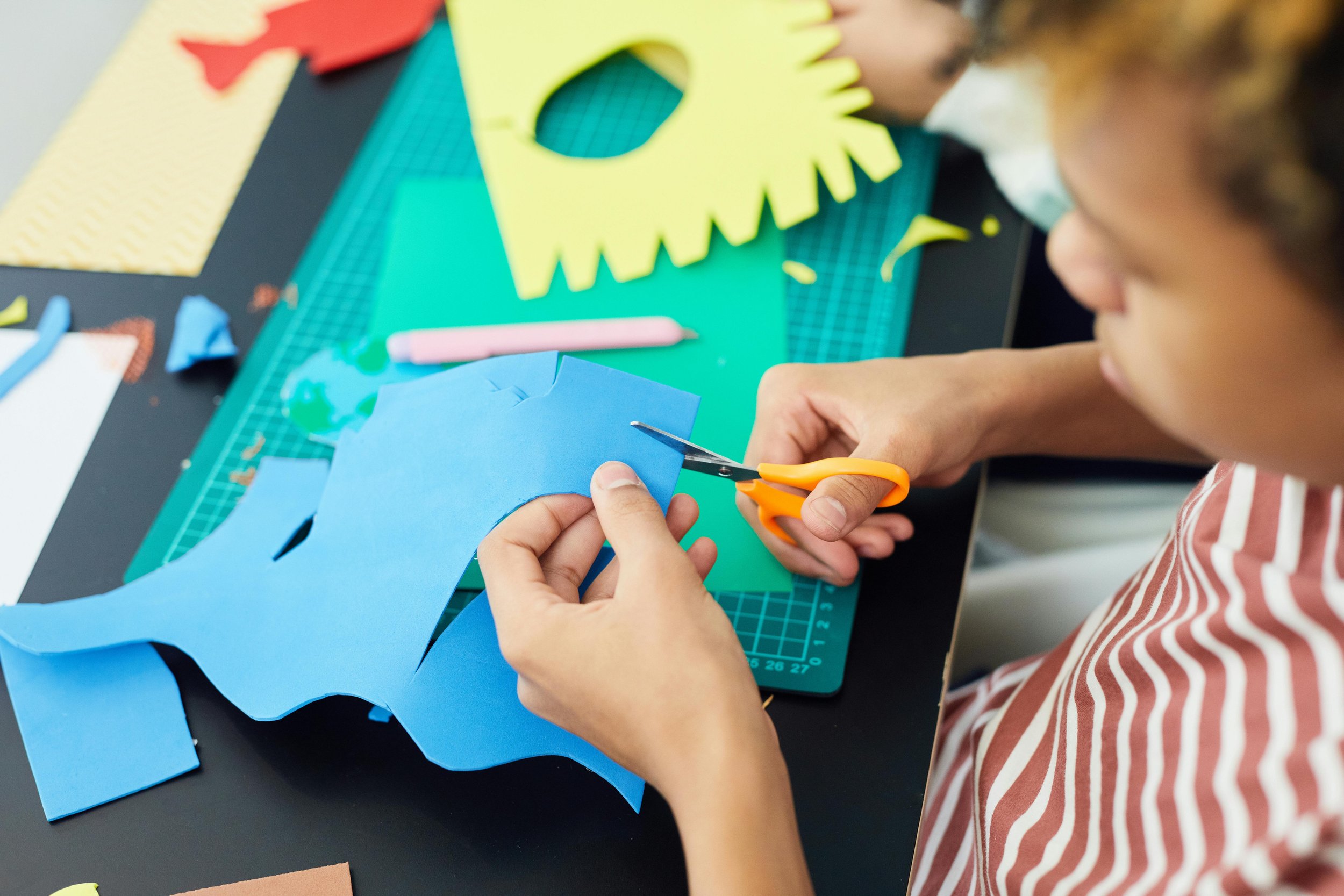 A person using orange scissors to cut blue paper in an arts and crafts project on a green cutting mat, with various colored paper pieces and craft tools around.