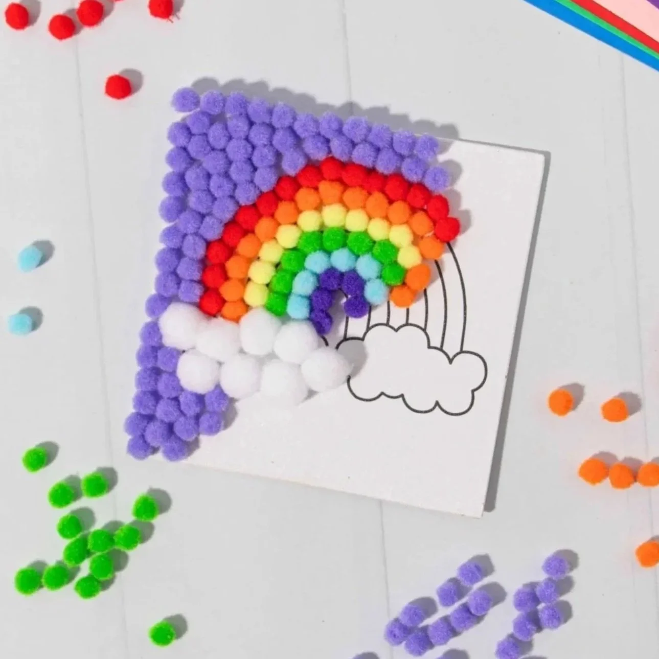Colorful pom-poms arranged as a rainbow on a drawing of a rainbow with clouds, on a white surface scattered with additional pom-poms.