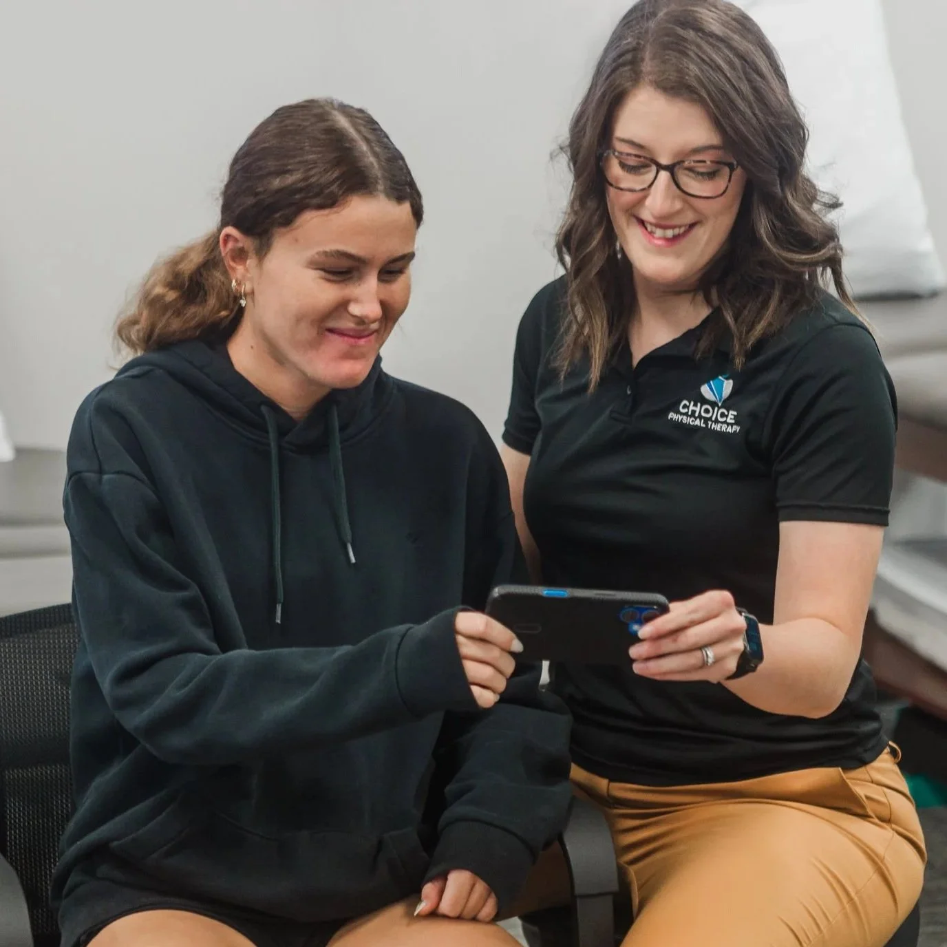 A woman with curly hair watching a video call with a smiling female doctor on her laptop in her home, sitting at a table with a mug in her hand.