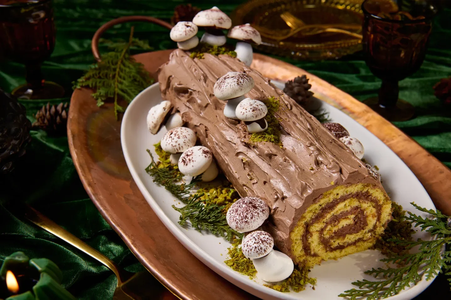 A chocolate yule log cake with mushroom decorations on top, served on a large white platter surrounded by greenery and festive table setting.
