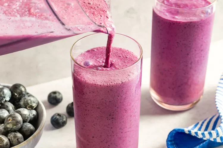 A glass of pink blueberry smoothie being poured from a blender, with a bowl of blueberries nearby and another glass of smoothie in the background.