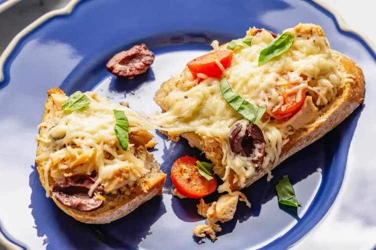 Close-up of an open-faced chicken and vegetable casserole on a slice of bread, topped with melted cheese, cherry tomatoes, olives, and fresh green herbs, served on a blue plate.