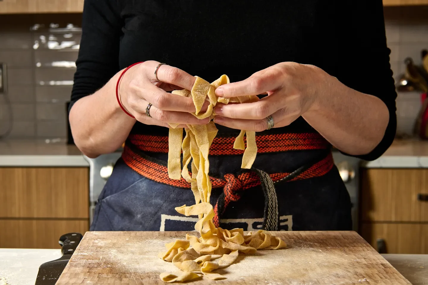 Person in a black shirt and an apron with a red and black belt making homemade pappardelle pasta, handling fresh pasta ribbons over a wooden cutting board in a kitchen.