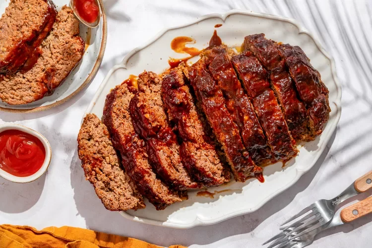 Barbecue meatloaf sliced on a white platter with barbecue sauce, with a side of ketchup and a small bowl of meatloaf slices.