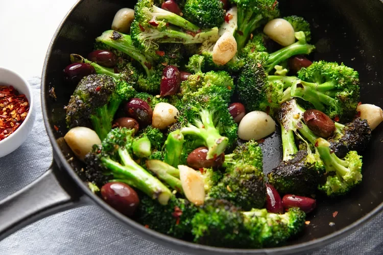 Sautéed broccoli with garlic and olives in a black skillet, with a small bowl of red pepper flakes nearby.