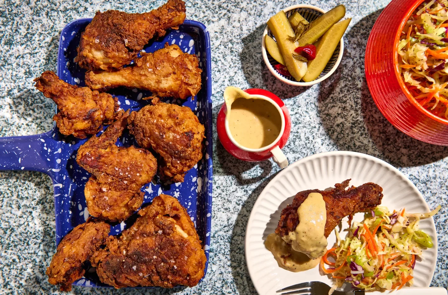 A plate of fried chicken pieces, a bowl of coleslaw, pickles, chicken with gravy, and a glass of iced tea on a speckled granite countertop.