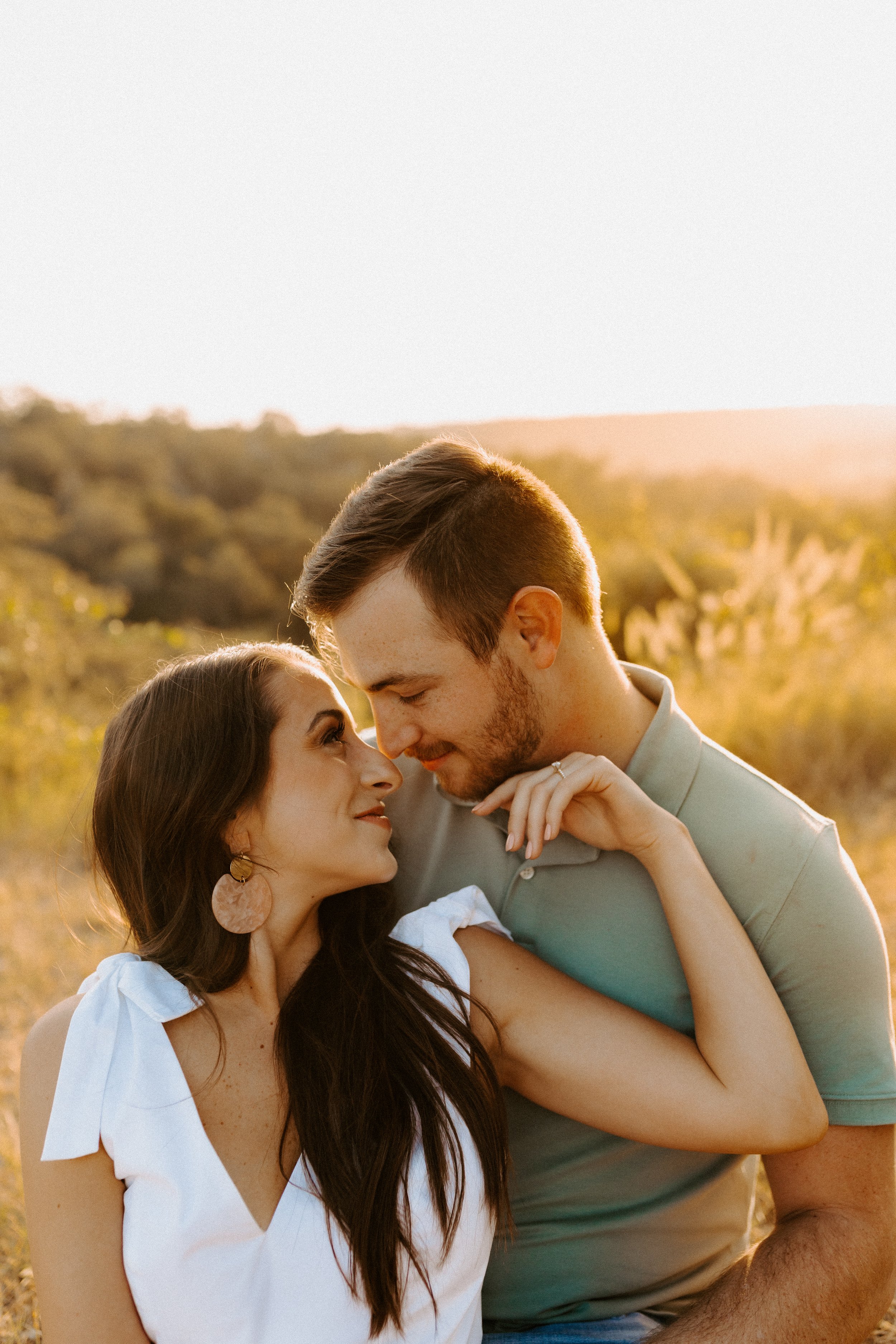 A couple sitting outdoors, gazing at each other affectionately during a golden hour sunset with a natural background.