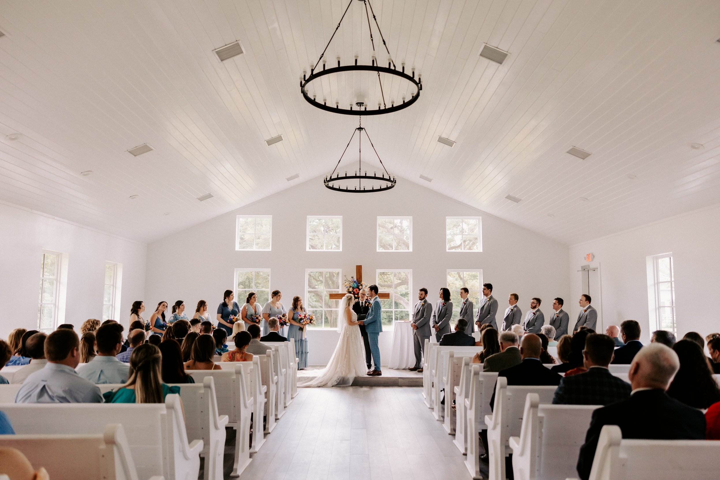 Wedding ceremony inside a chapel with bride and groom at the altar, bridesmaids and groomsmen standing on either side, and guests seated in white pews.