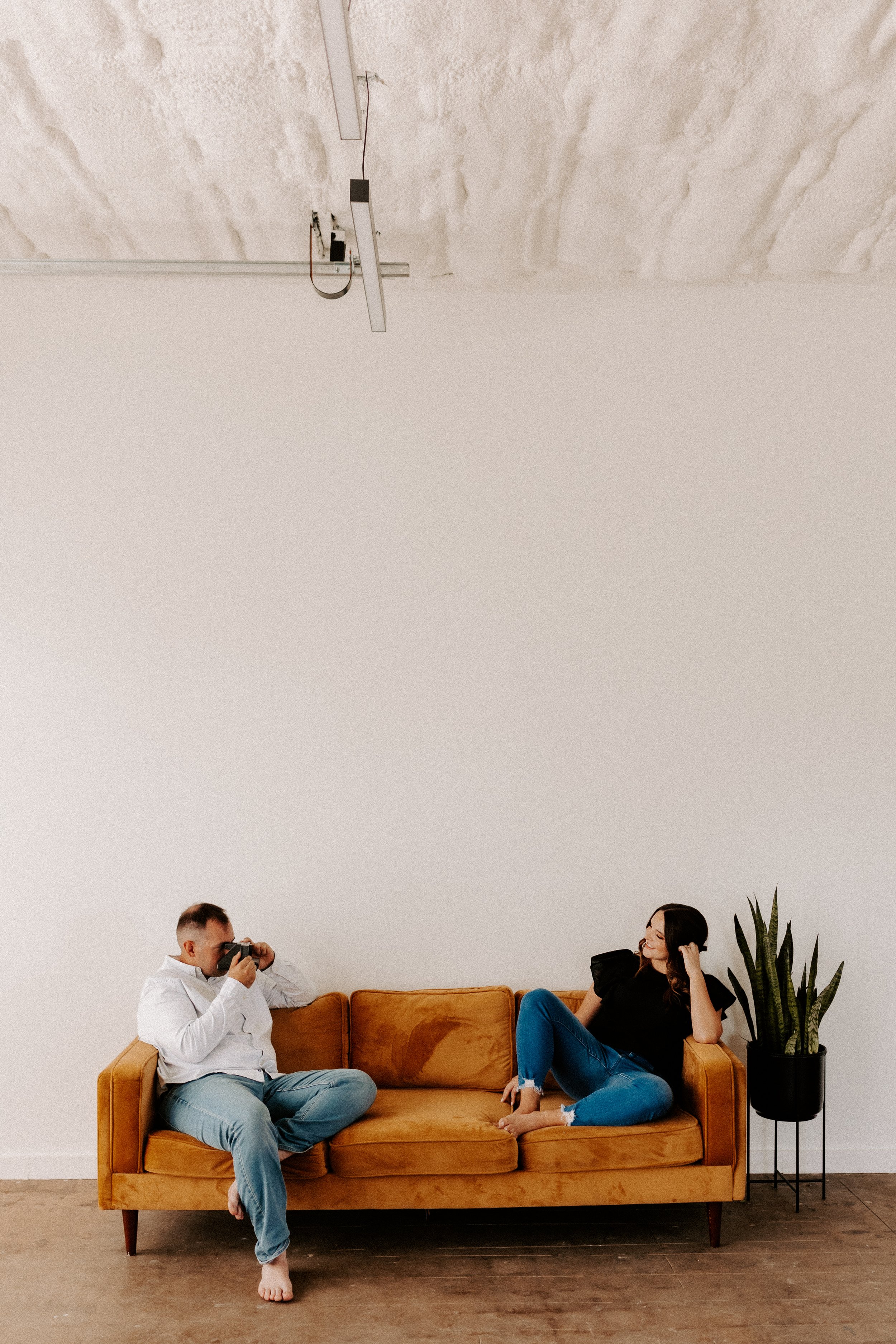 Two people sitting on an orange couch; one person is using a camera and the other is sitting with a potted plant nearby.