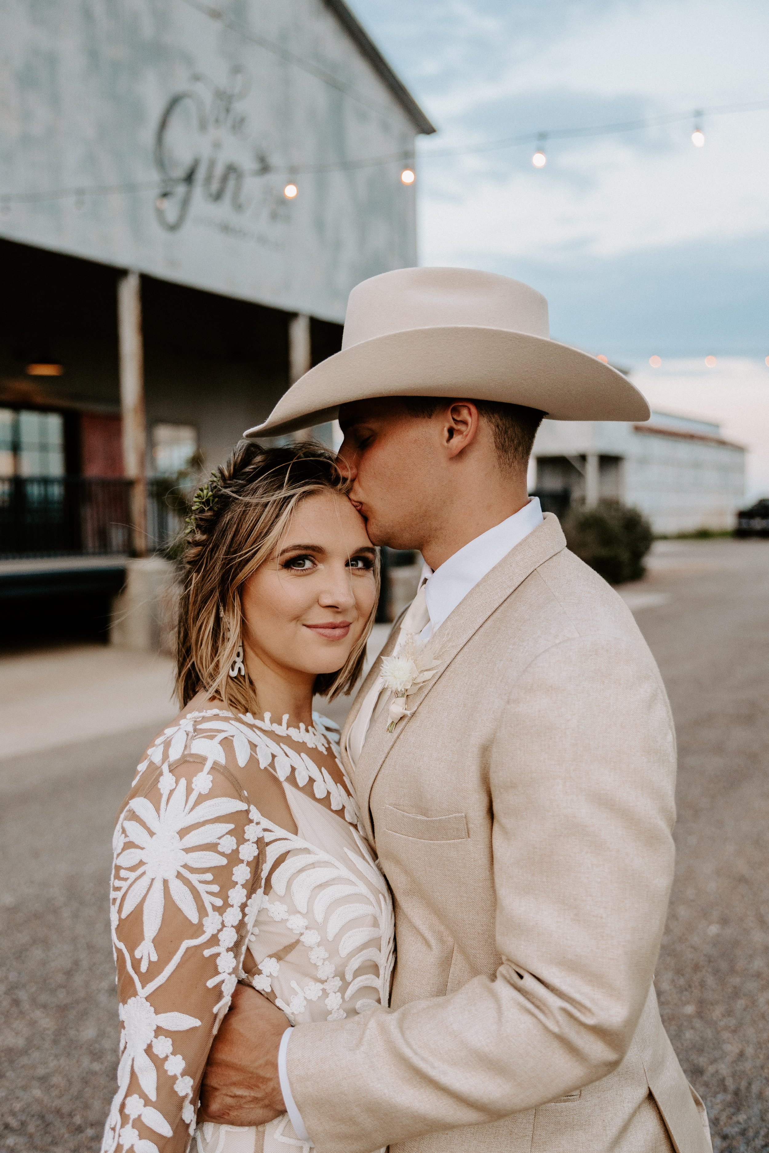 Couple embracing at a rustic outdoor venue, man in cowboy hat kissing woman's forehead, woman wearing a floral lace dress.