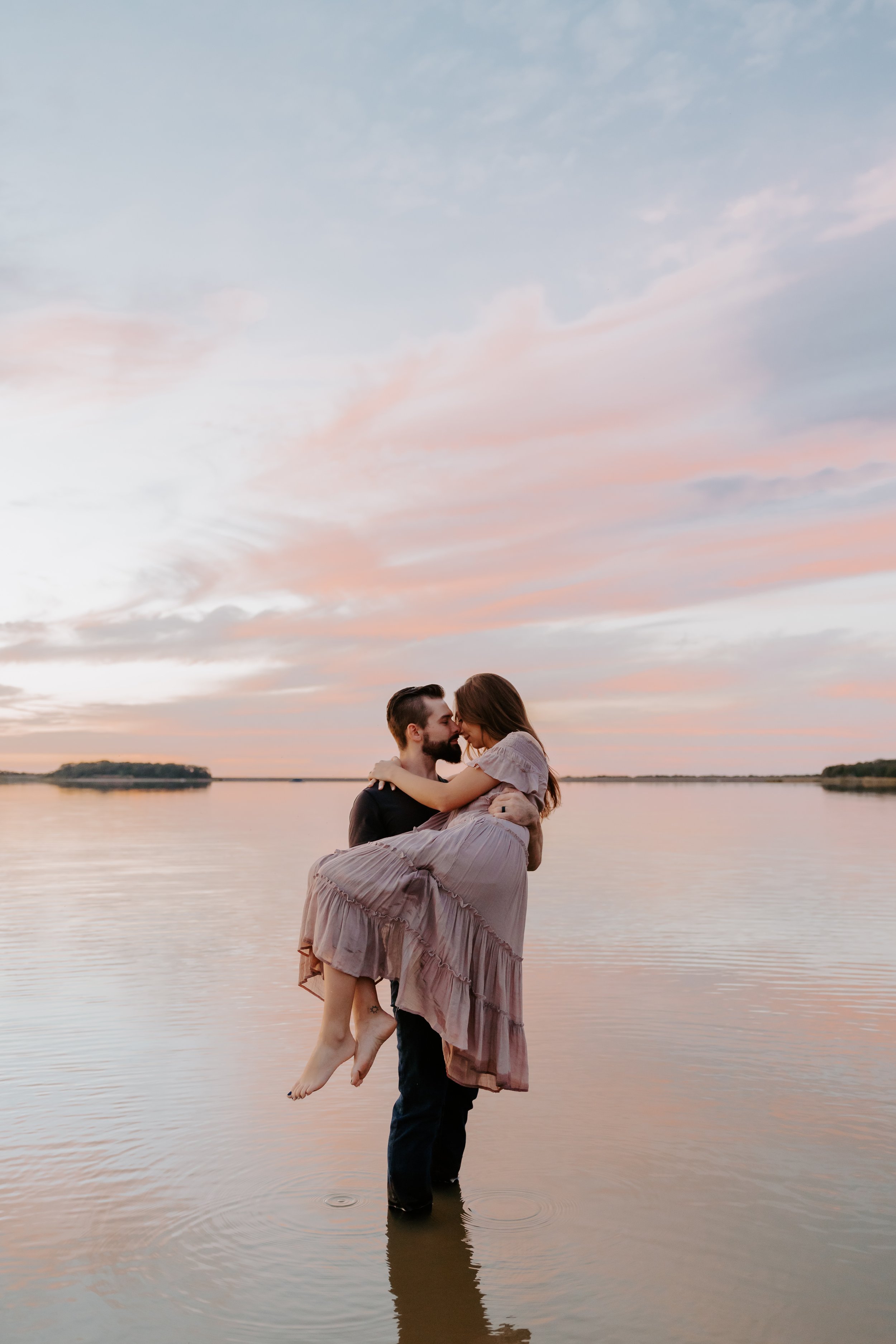 A romantic couple by a lake at sunset, with a man holding a woman in his arms, surrounded by pink and orange clouds.