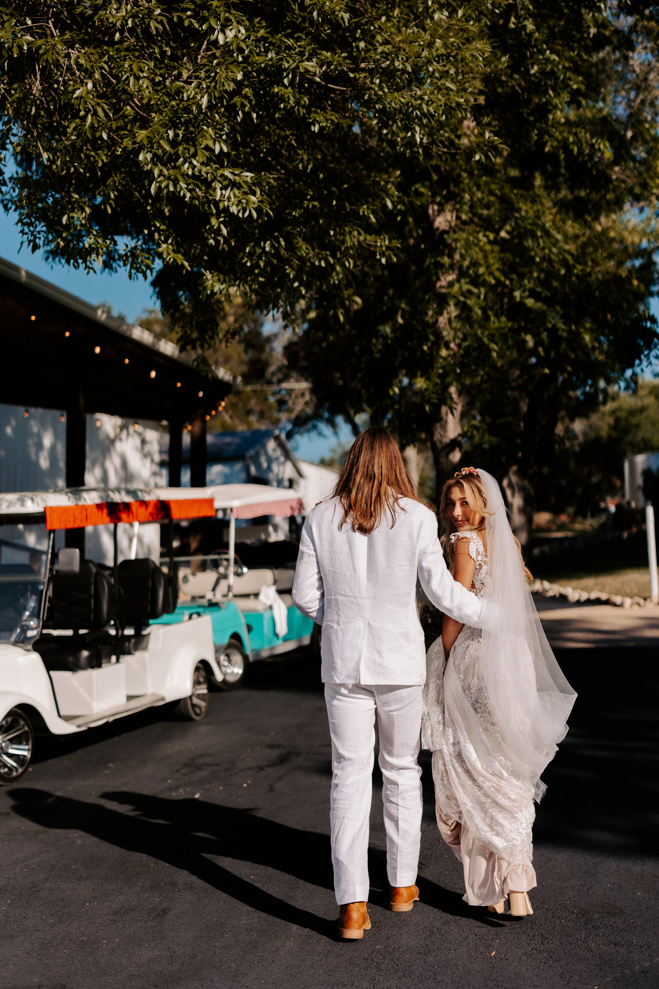 Bride and groom walking outdoors, the groom in a white suit and the bride in a lace wedding dress with a veil, near golf carts under trees.
