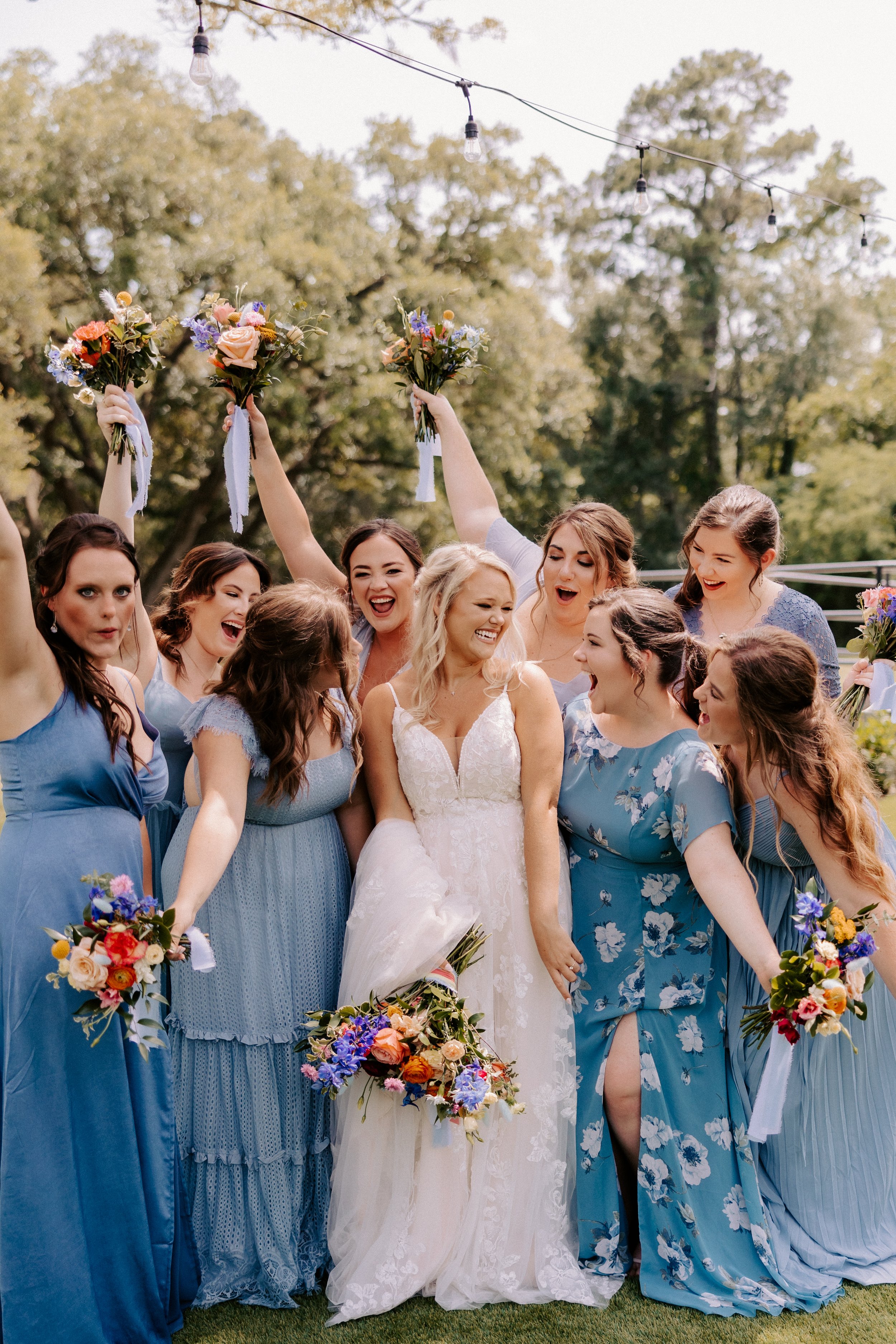 Bride and bridesmaids in blue dresses holding bouquets, celebrating outdoors.