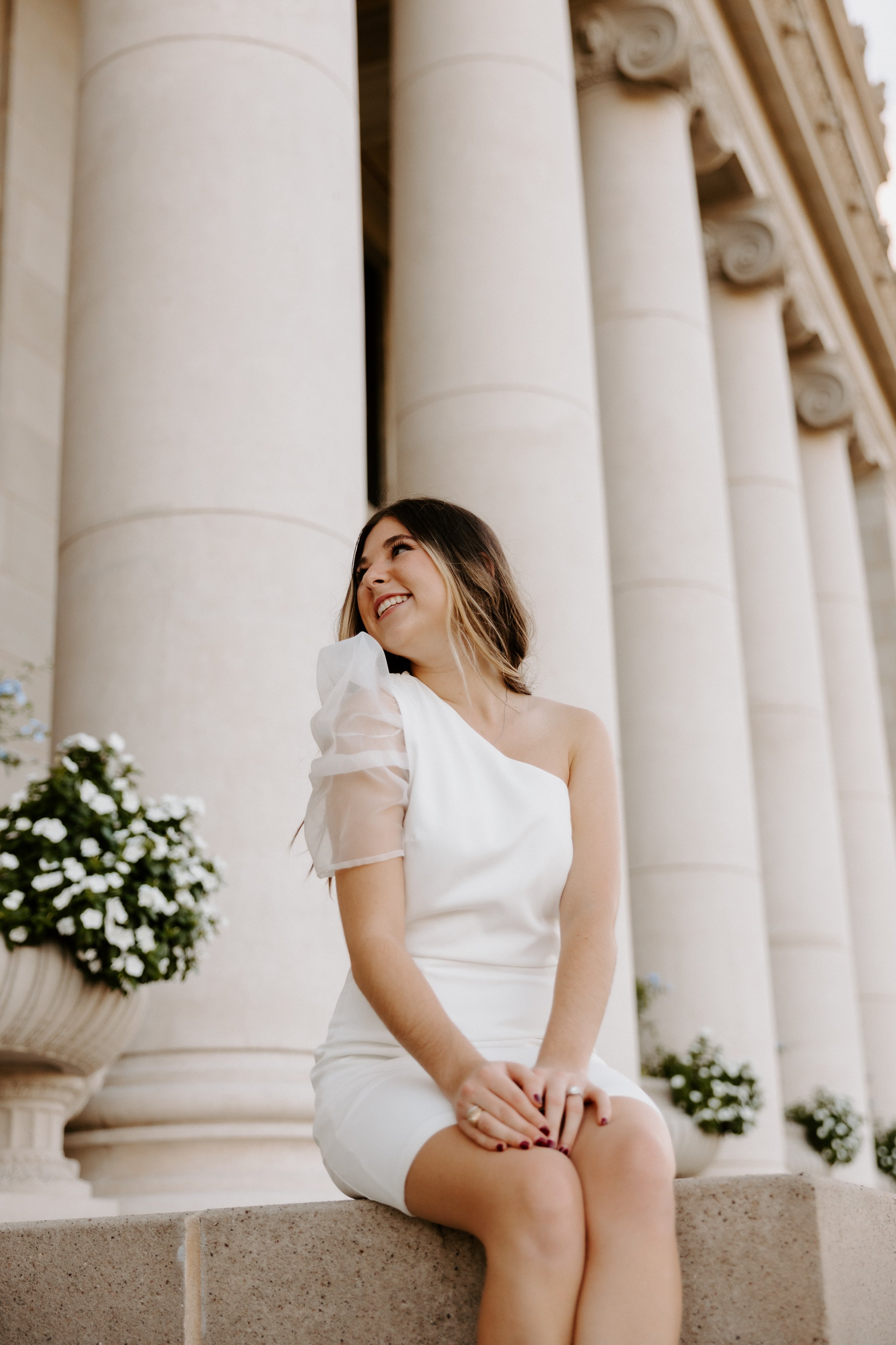 Woman in white dress sitting on stone ledge with large columns in background.