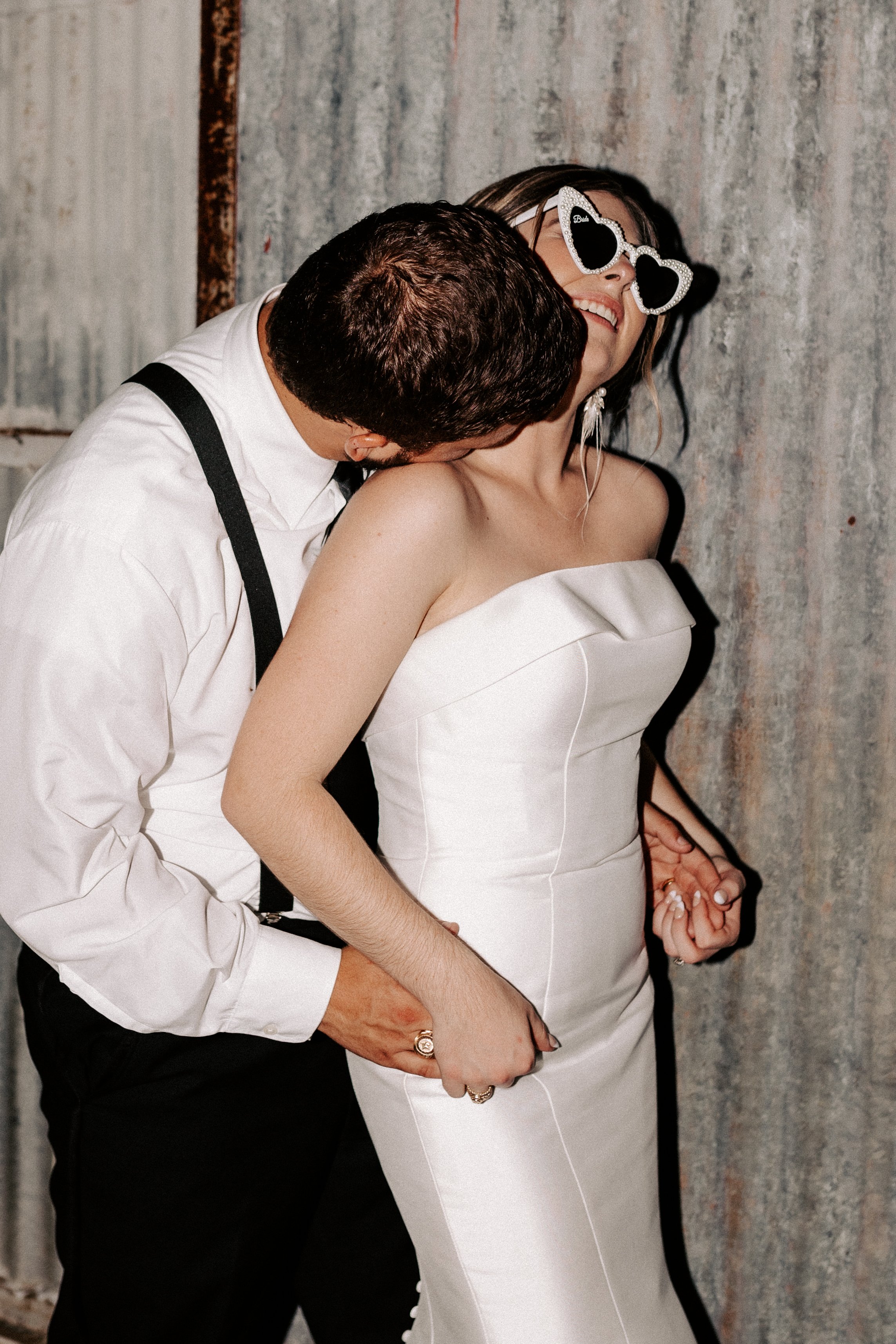 A couple standing close together, with the man wearing a white shirt and black suspenders, embracing a woman in a strapless white dress. The woman is wearing heart-shaped sunglasses and smiling.