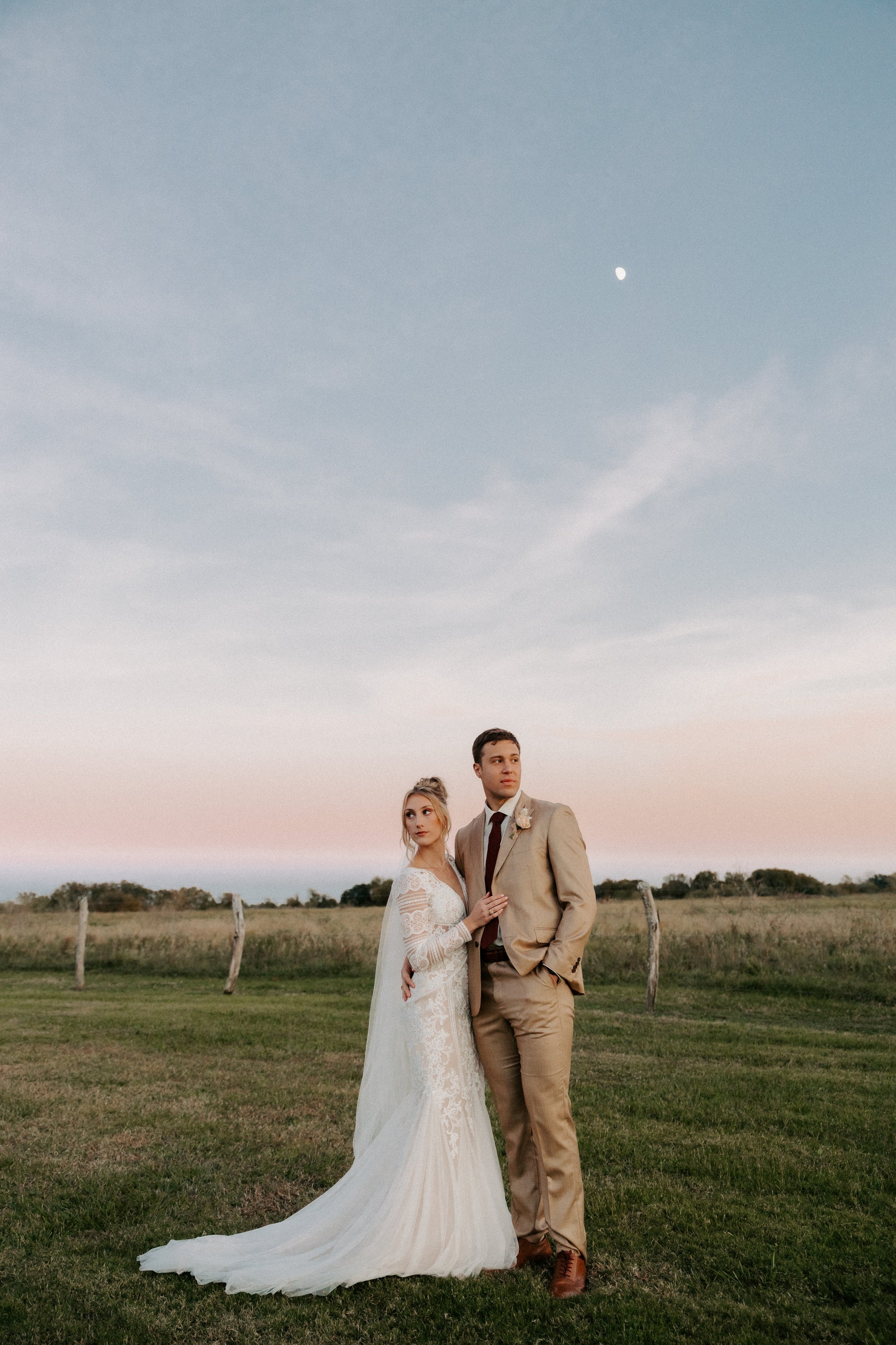 Bride in a white lace wedding dress and groom in a tan suit standing in a field under a clear sky with the moon visible.