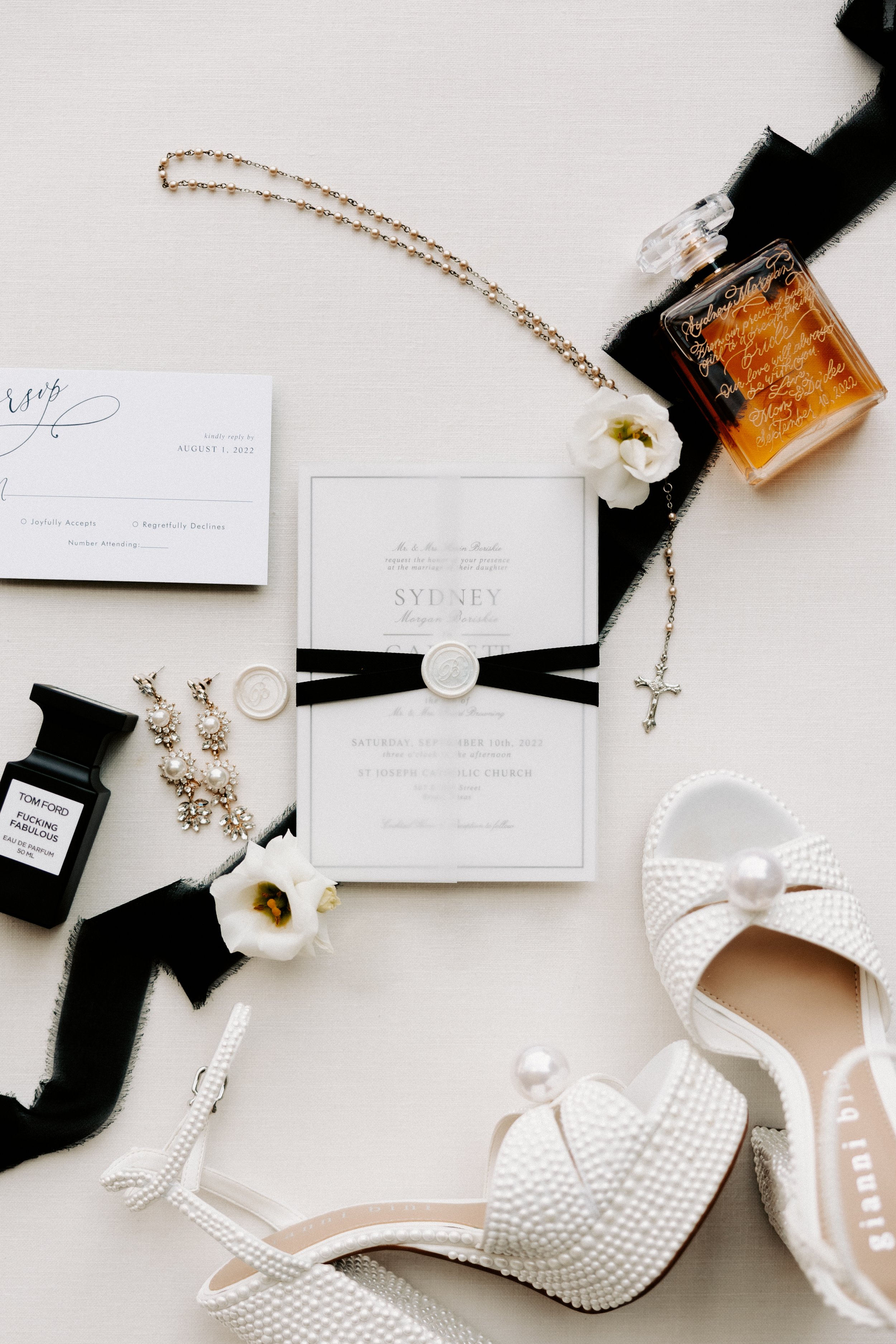 Flat lay of wedding items including invitations, RSVP card, perfume bottles, pearl earrings, white sandals, a rosary, and white flowers on a light background.