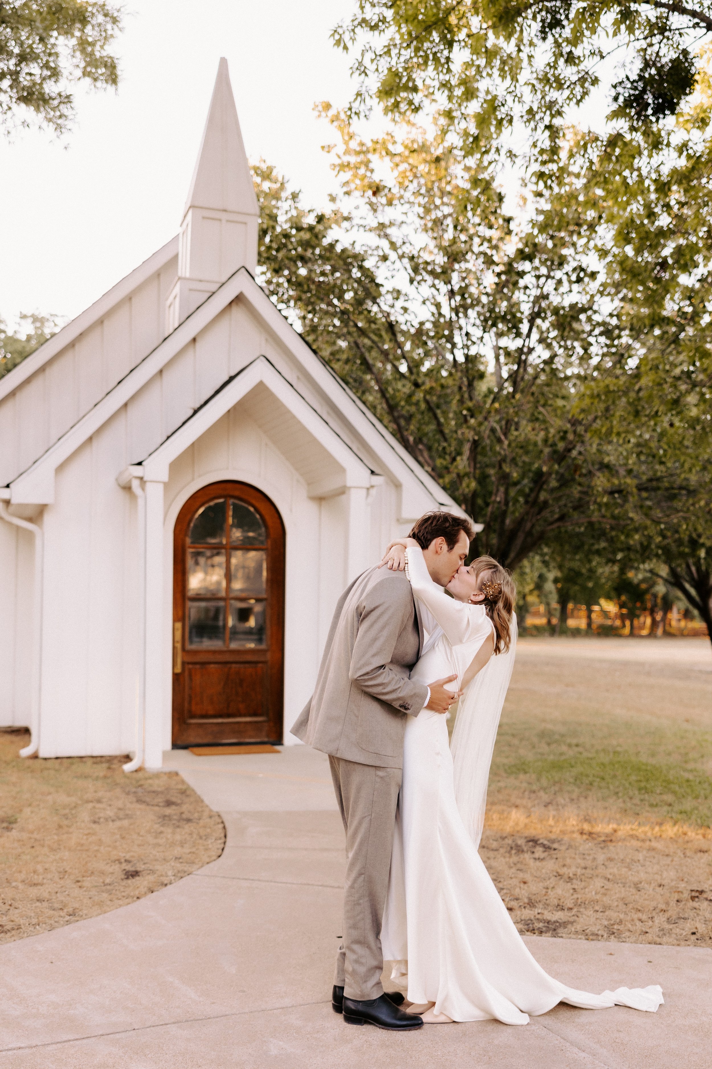 A couple sharing a kiss in front of a small white chapel with trees in the background. The woman is wearing a white wedding dress and the man is in a light-colored suit.