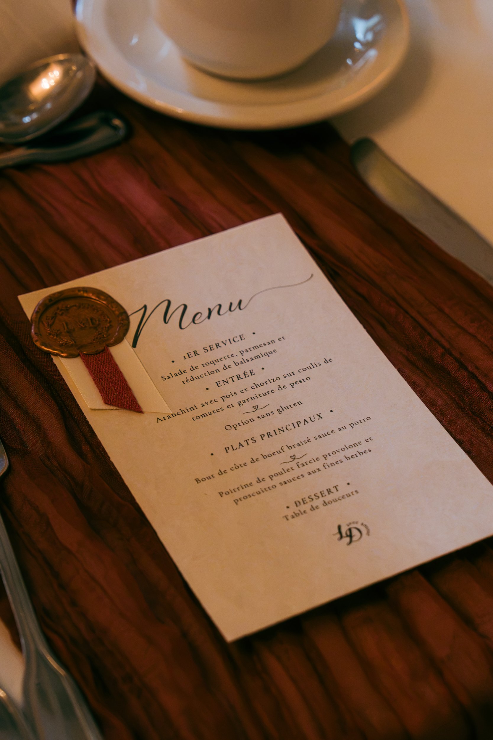 Un menu de restaurant en français avec décoration en cire rougeâtre et une tasse blanche sur une assiette. Le menu est placé sur une table en bois.