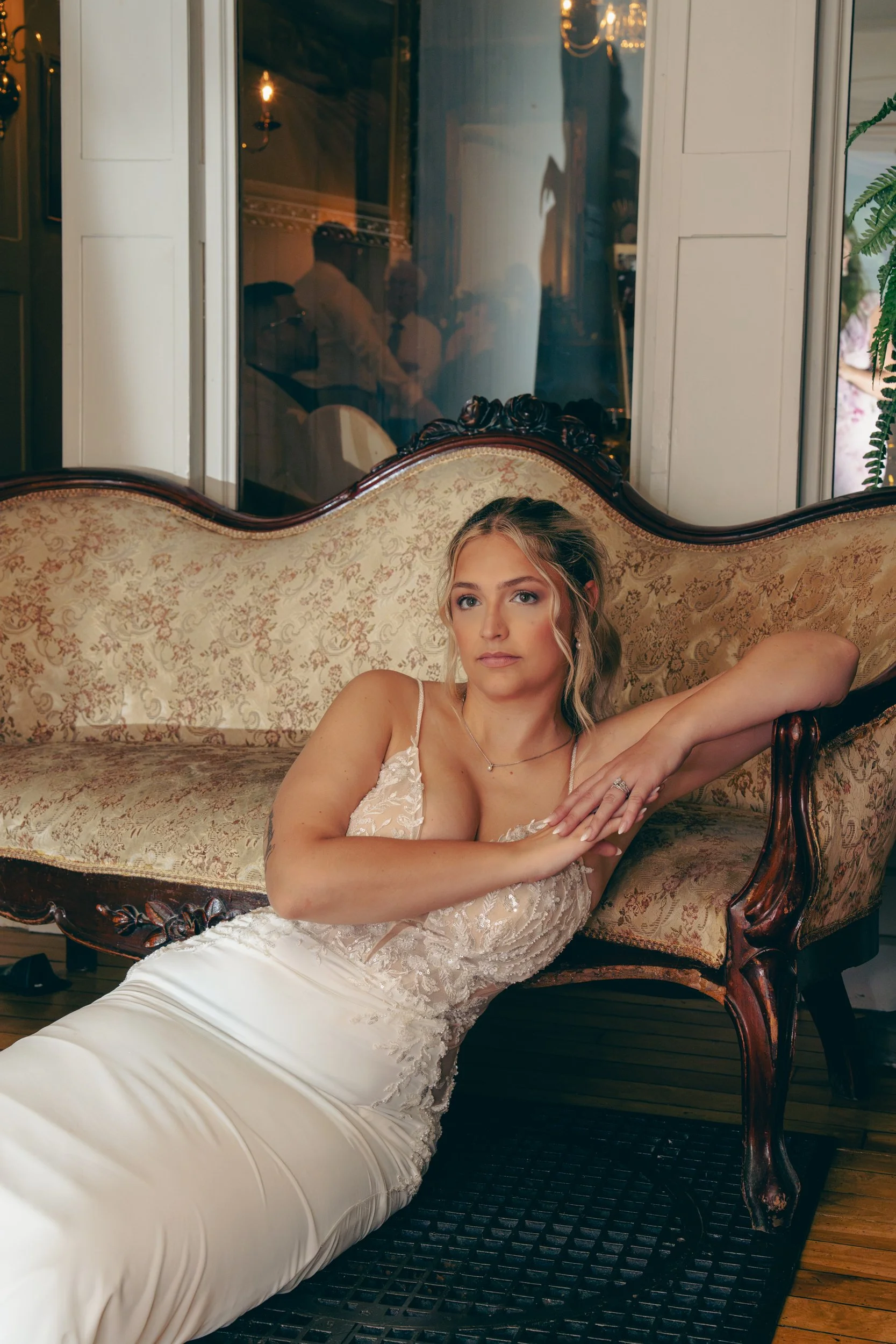 Une femme en robe de mariage blanche repose sur un canapé en bois sculpté avec un tissus à motif floral, à l'intérieur d'une pièce élégante avec des miroirs et un éclairage tamisé.