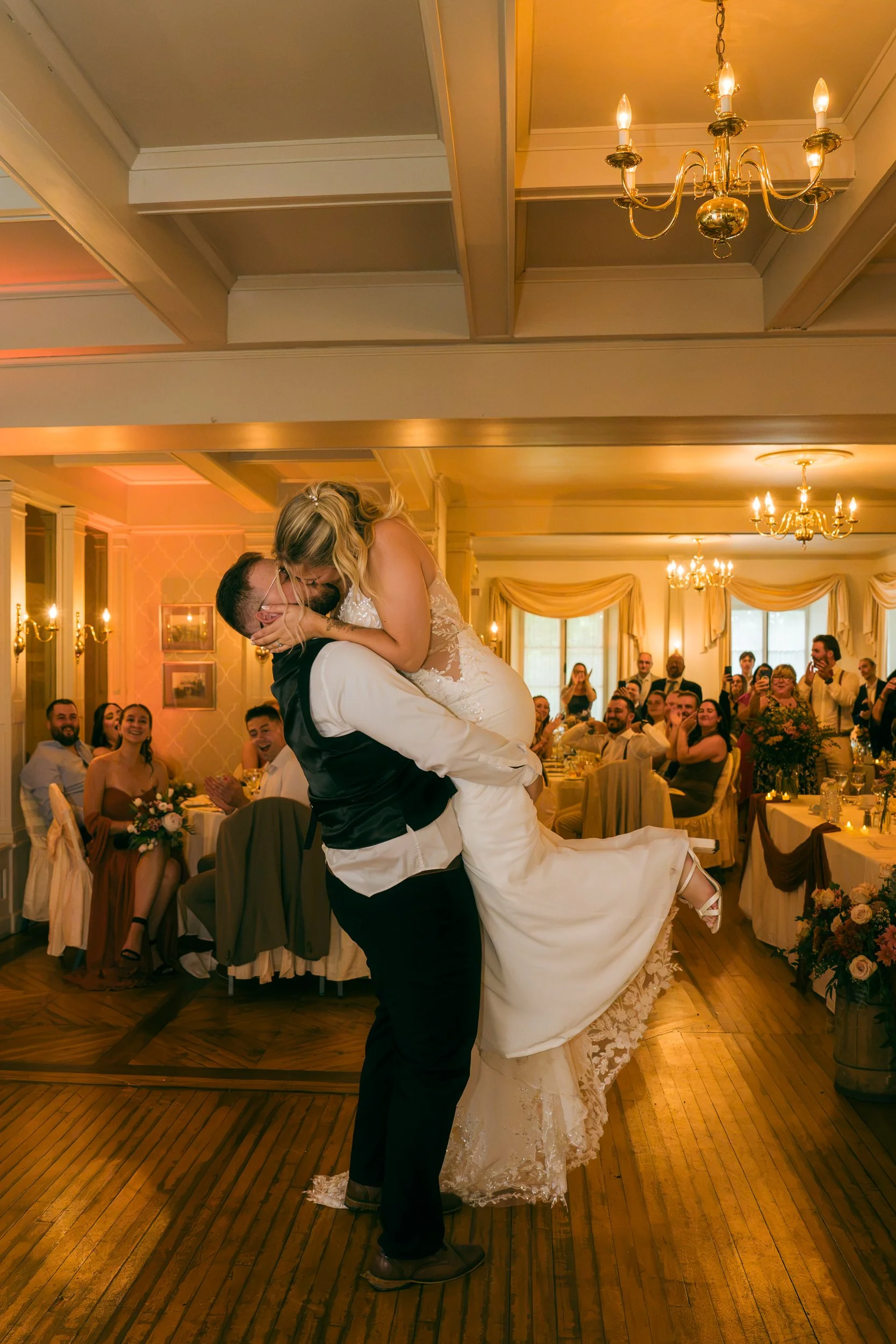 Un couple de mariés en pleine danse lors d'un mariage, entouré d'invités assis à des tables décorées, dans une salle élégante avec des chandeliers en or et des fenêtres avec rideaux beige.