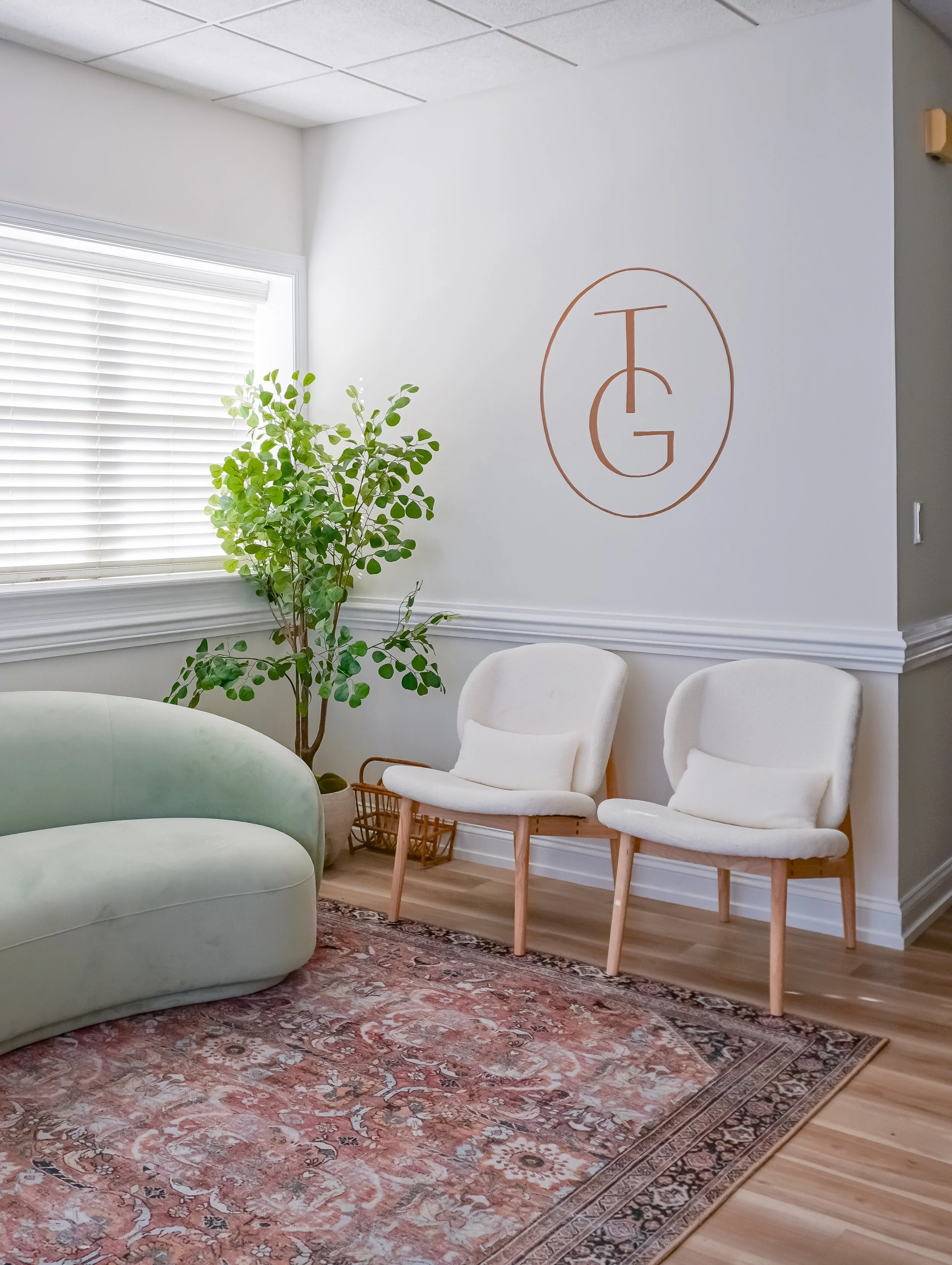 Bright living room with a green sofa, two white armchairs, a leafy indoor plant, patterned area rug, window blinds, and monogram wall art.