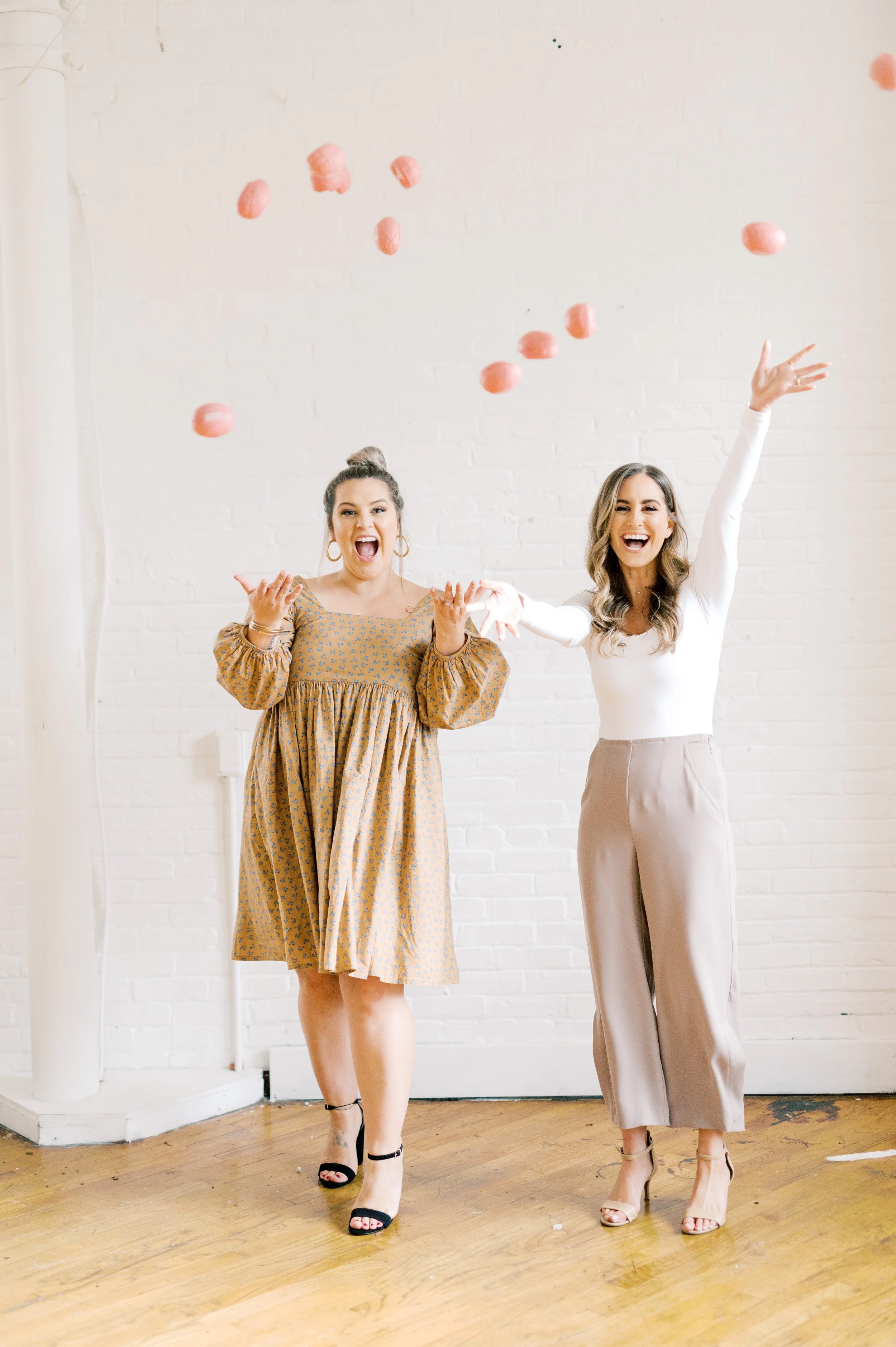 Em and Jen, throwing ShrinkChicks Brains in the air in front of a white brick wall.