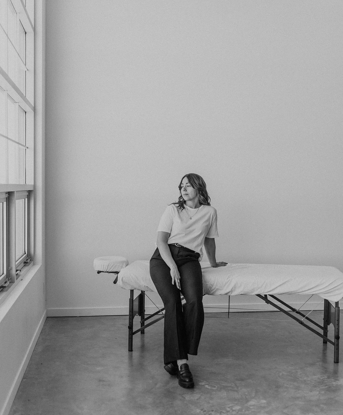 A woman sitting on the edge of a bed in a minimalist room with large windows and plain walls, dressed in a t-shirt and black pants.