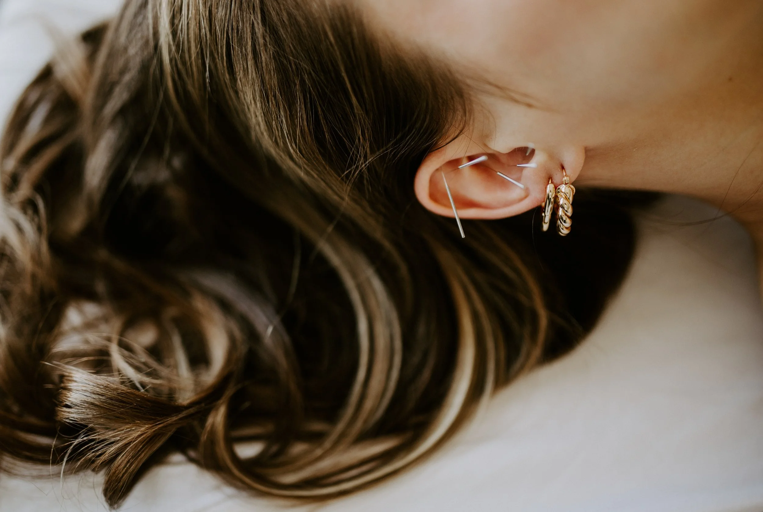 Close-up of a woman's head with detailed view of brown wavy hair and multiple earrings including a hoop and small studs.