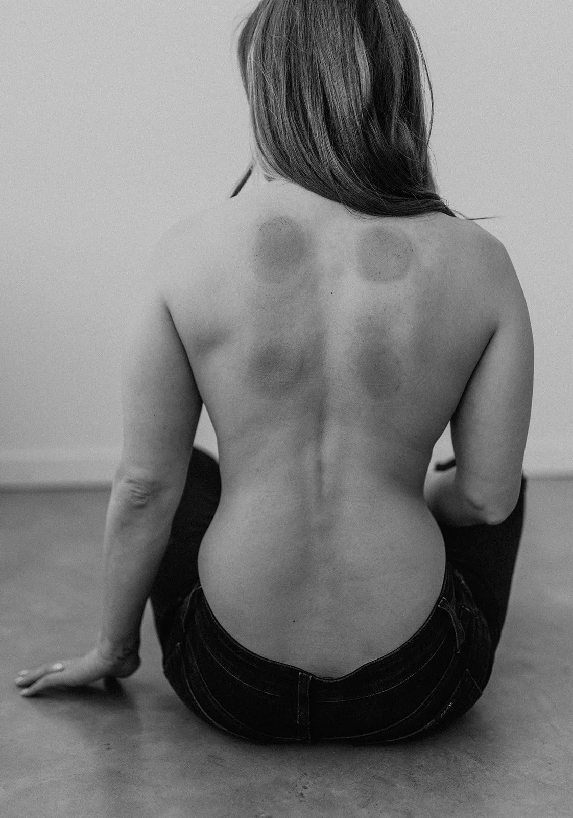 Black and white photograph of a woman sitting on the floor with her back to the camera. She is topless, with visible shoulder blades and a slender back, wearing dark jeans. Her hair is shoulder-length, and she is holding a pencil in her right hand.