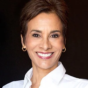 Person smiling with short hair and white blouse in front of a dark background.